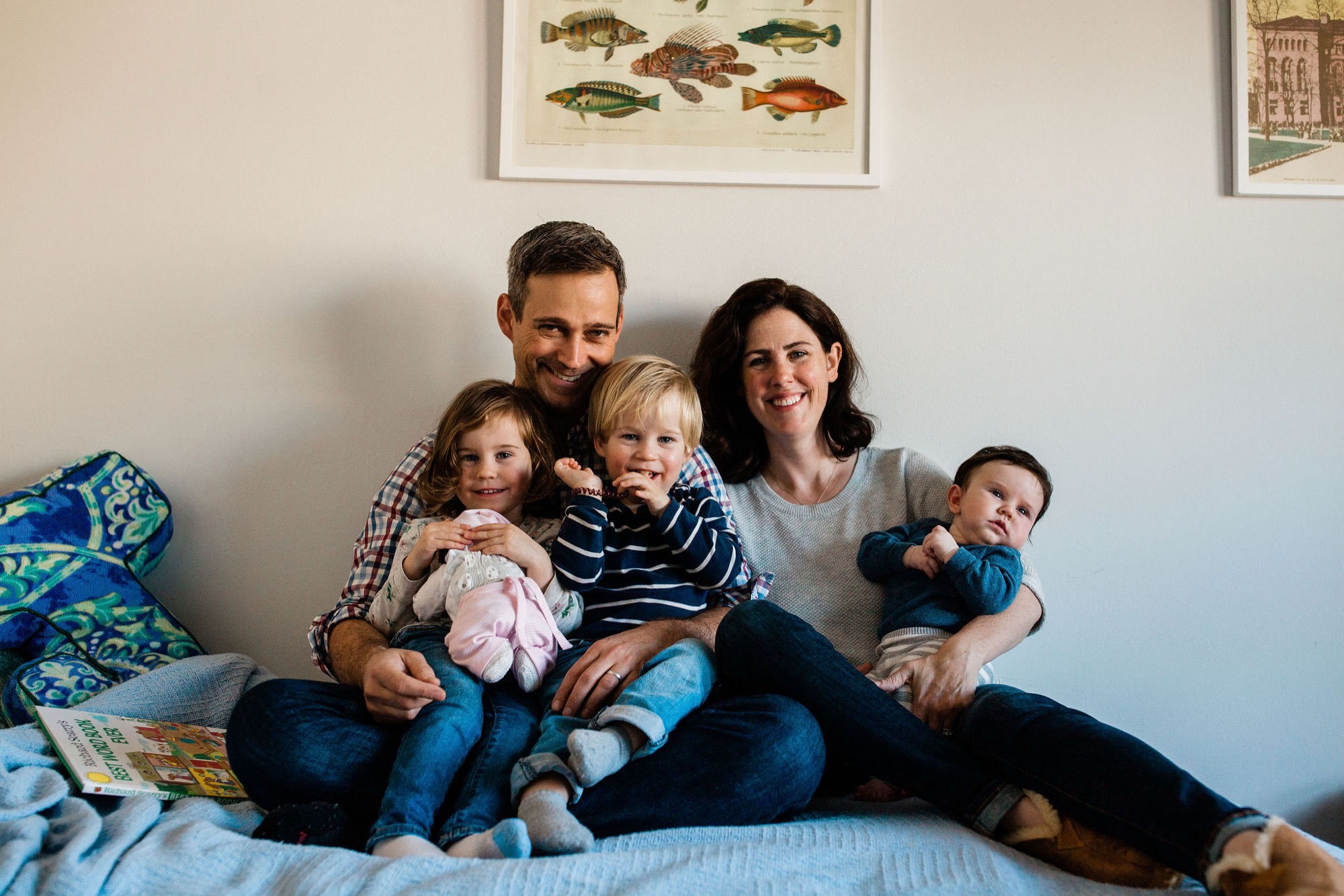 Family of five sits together on a bed, the parents smiling warmly while their three young children lean into them, bathed in soft natural light