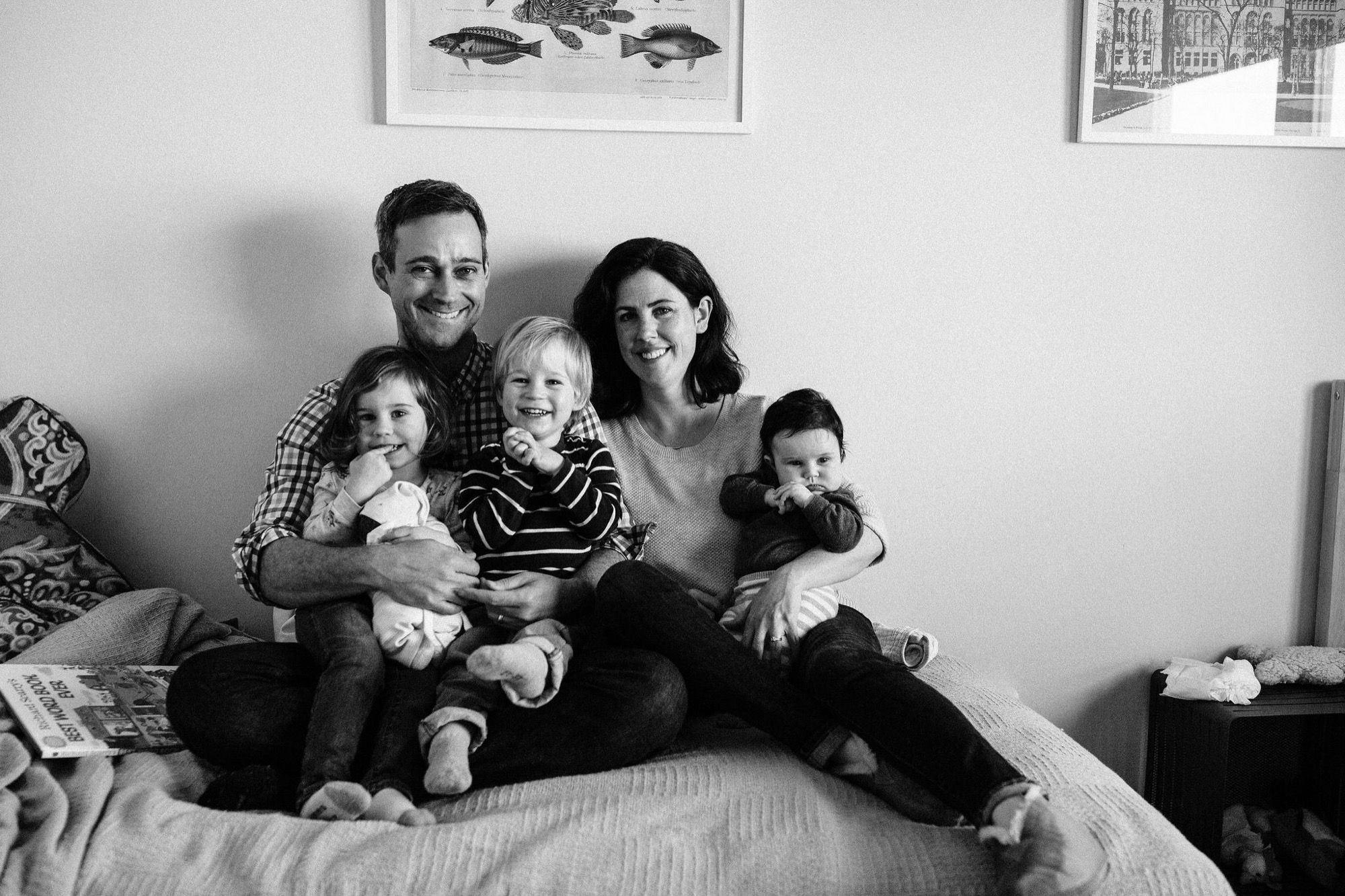 Parents and three young children sit together on a bed, all smiling genuinely at the camera in soft natural light