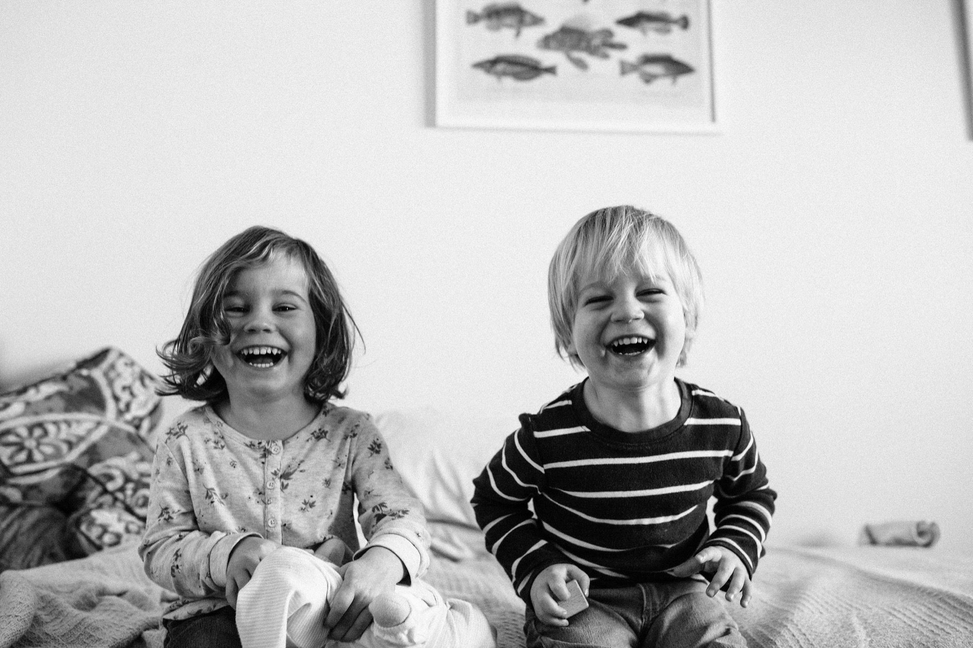 Two young children laugh freely on a bed, their genuine joy captured in black and white against a bright, minimalist bedroom wall