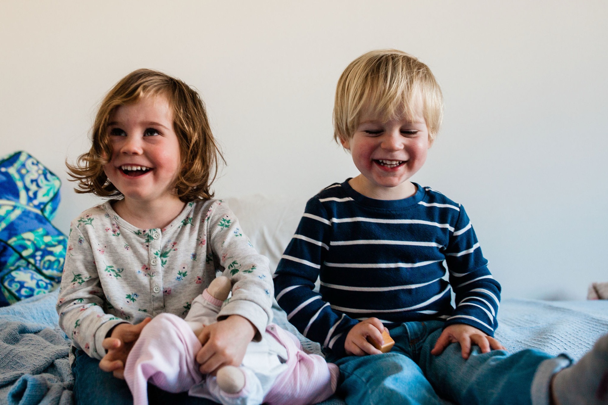 Two young children sit together on a bed, both laughing with genuine joy and delight, bathed in soft natural light against a neutral white wall