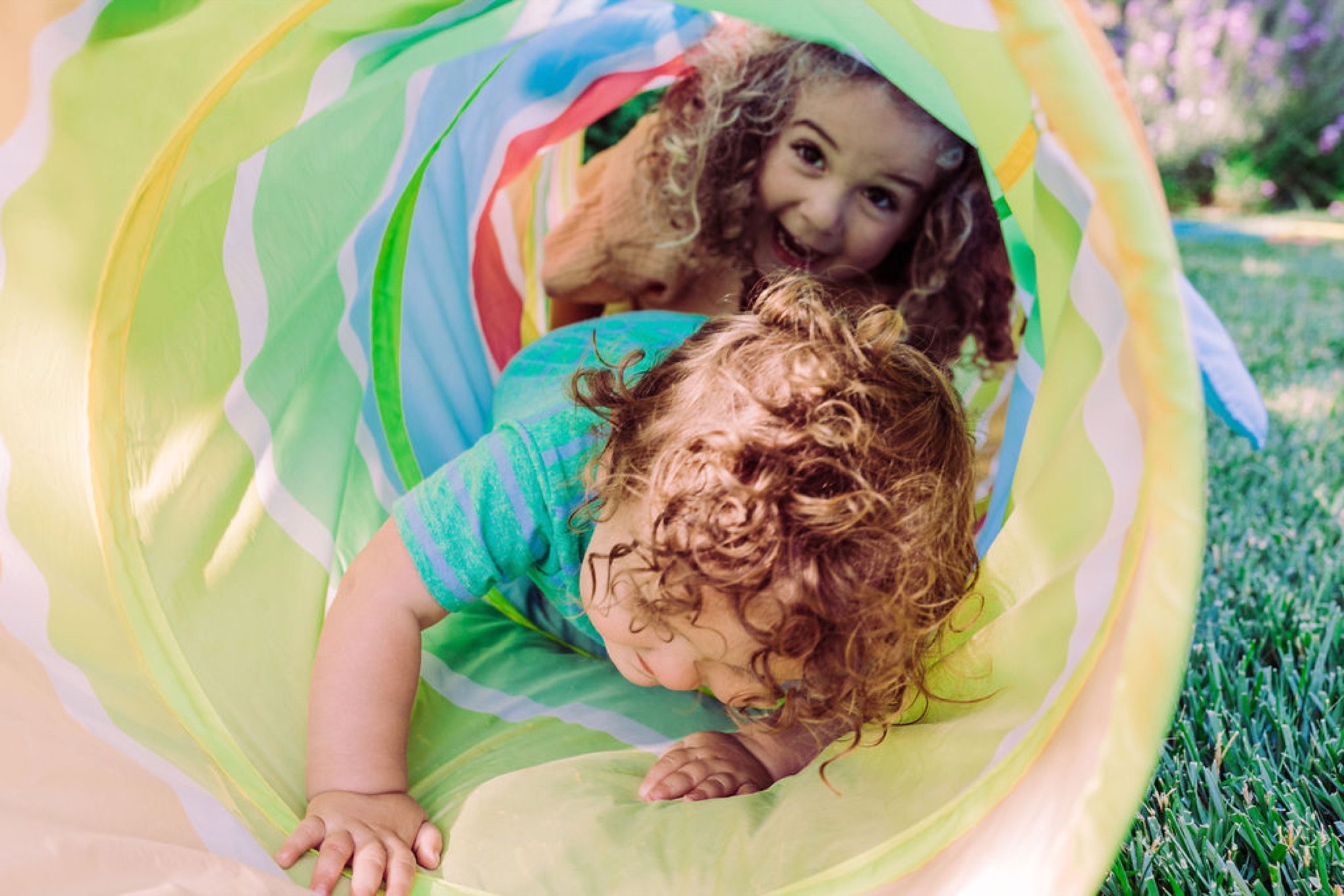 Two young children with curly hair crawl through a colorful fabric tunnel on grass, sharing joyful expressions and playful connection