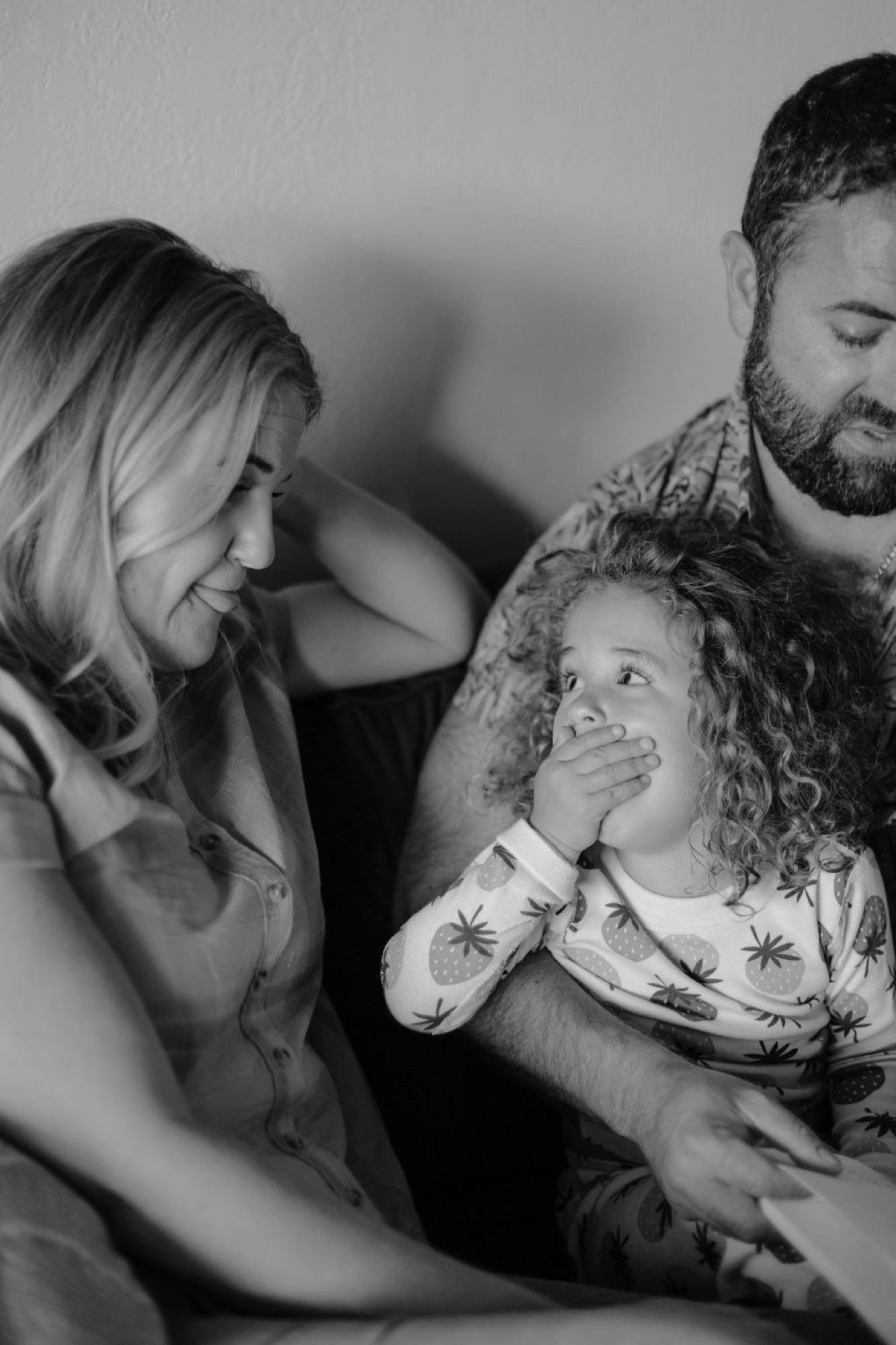A young girl with curly hair covers her mouth in delighted surprise while her parents lean in close, sharing an intimate moment of genuine family connection