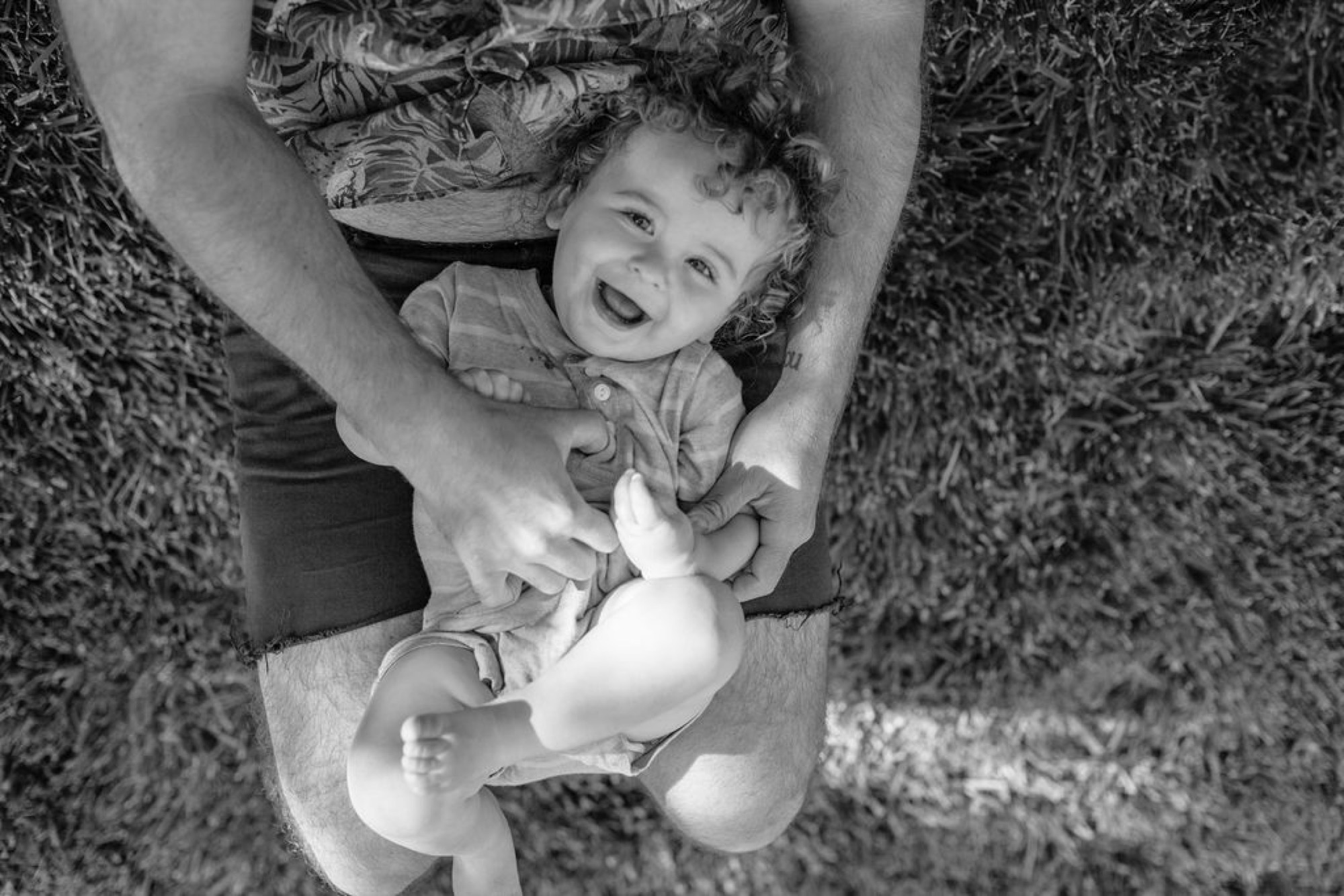 A toddler with curly hair beams with pure joy while being held overhead on grass, surrounded by textured foliage