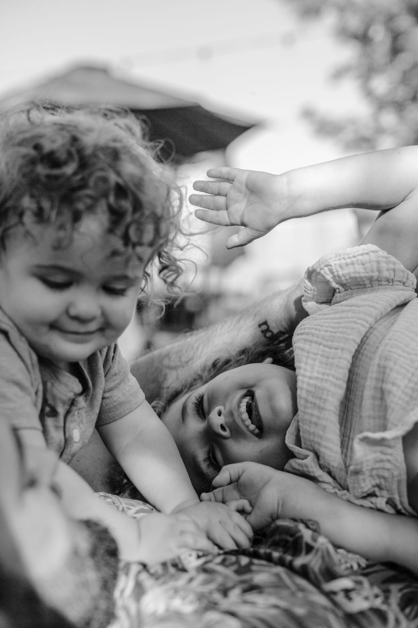 Two young children with curly hair smile and embrace on a patterned blanket, their genuine joy captured in soft, natural outdoor light