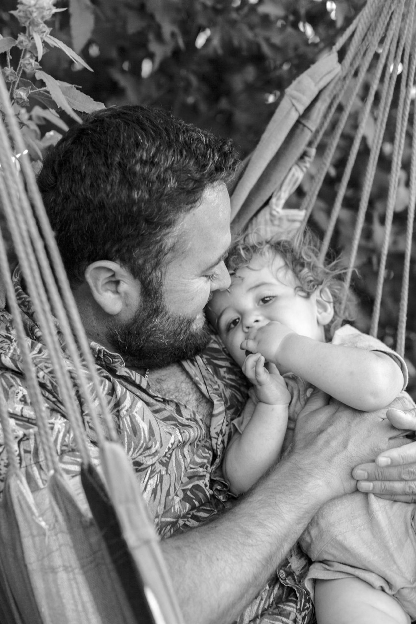 A bearded father gazes tenderly at his toddler while surrounded by palm fronds, the child's fingers in mouth as soft natural light illuminates their connection