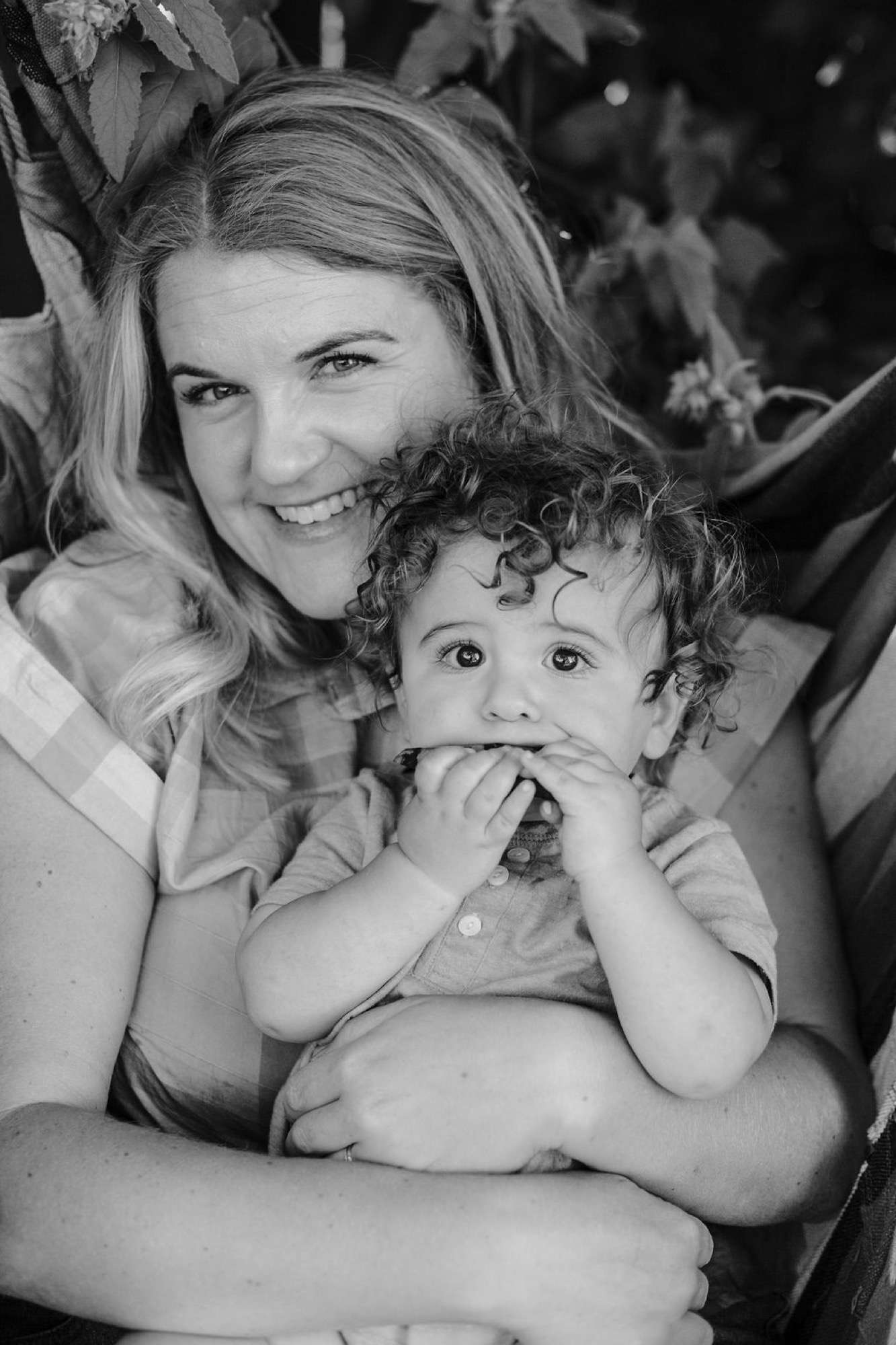 A woman beams at the camera while cradling her toddler, whose curly hair and wide eyes convey curious wonder against the soft garden backdrop