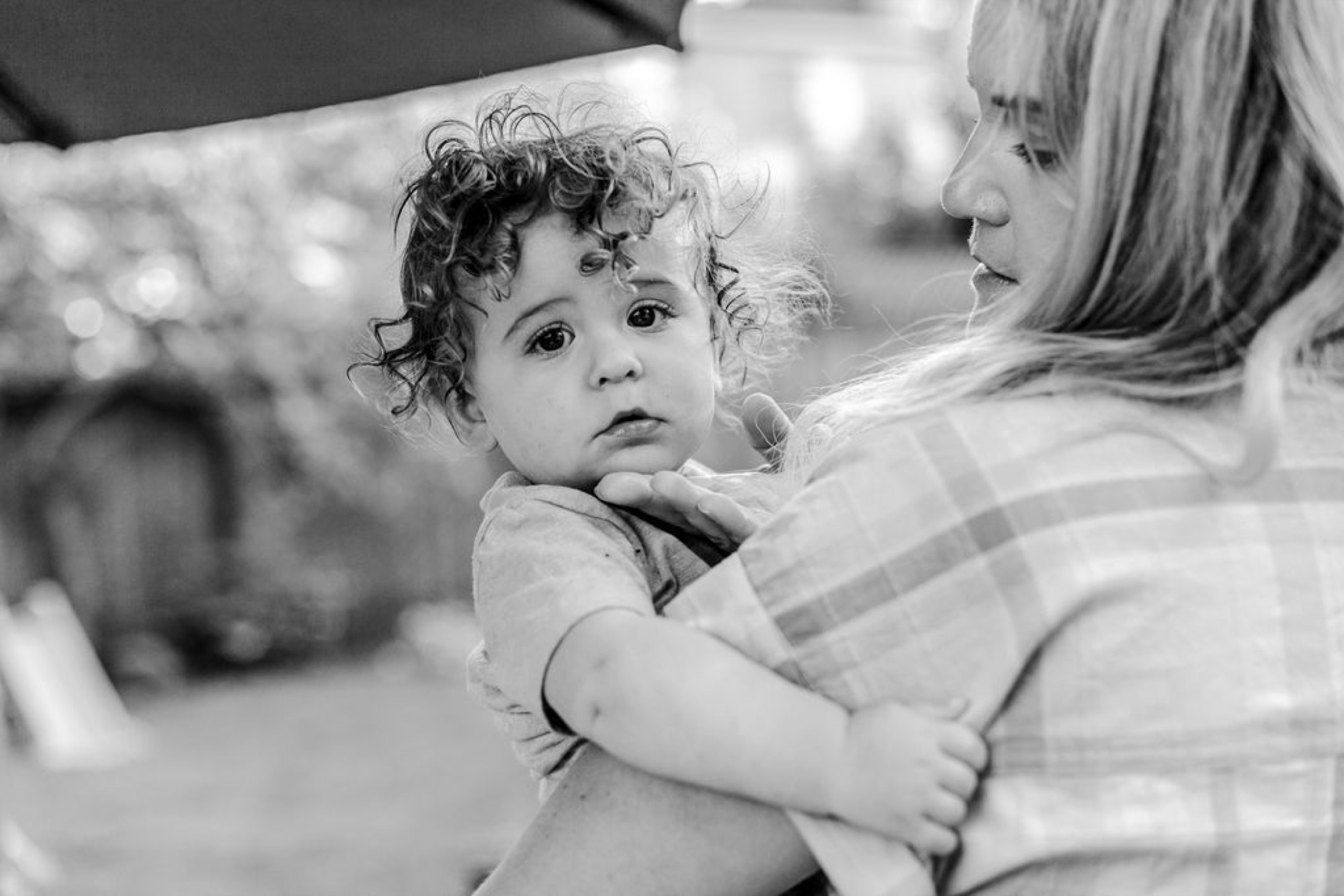 A toddler with curly hair gazes back over their shoulder while held by a woman in a striped shirt, captured in soft natural light