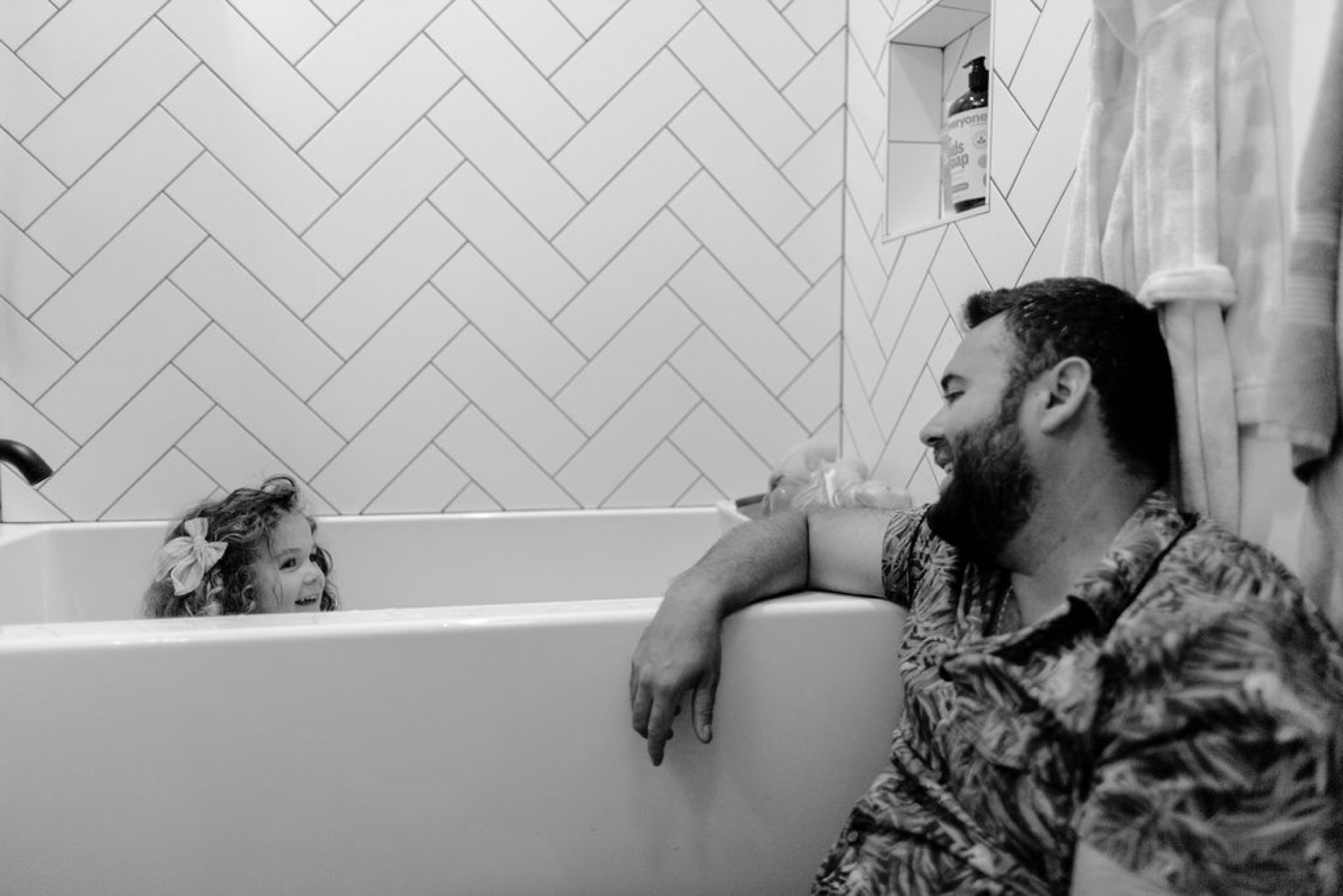 A bearded father and young child share a joyful bath-time moment, their genuine smiles and relaxed postures creating an intimate family connection in a bright, minimalist bathroom