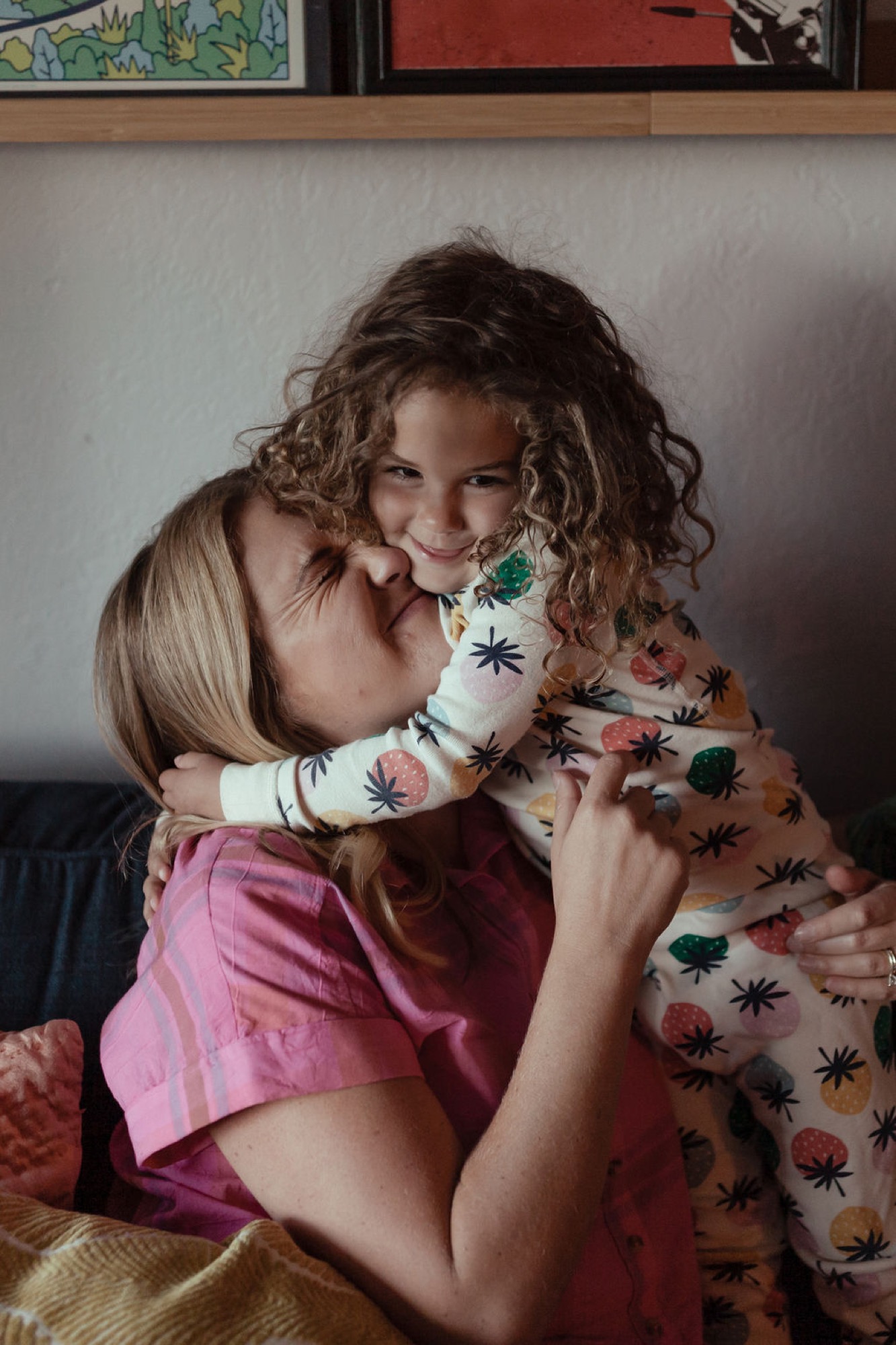 A mother tenderly kisses her young daughter's forehead while holding her close, both smiling warmly in soft indoor light