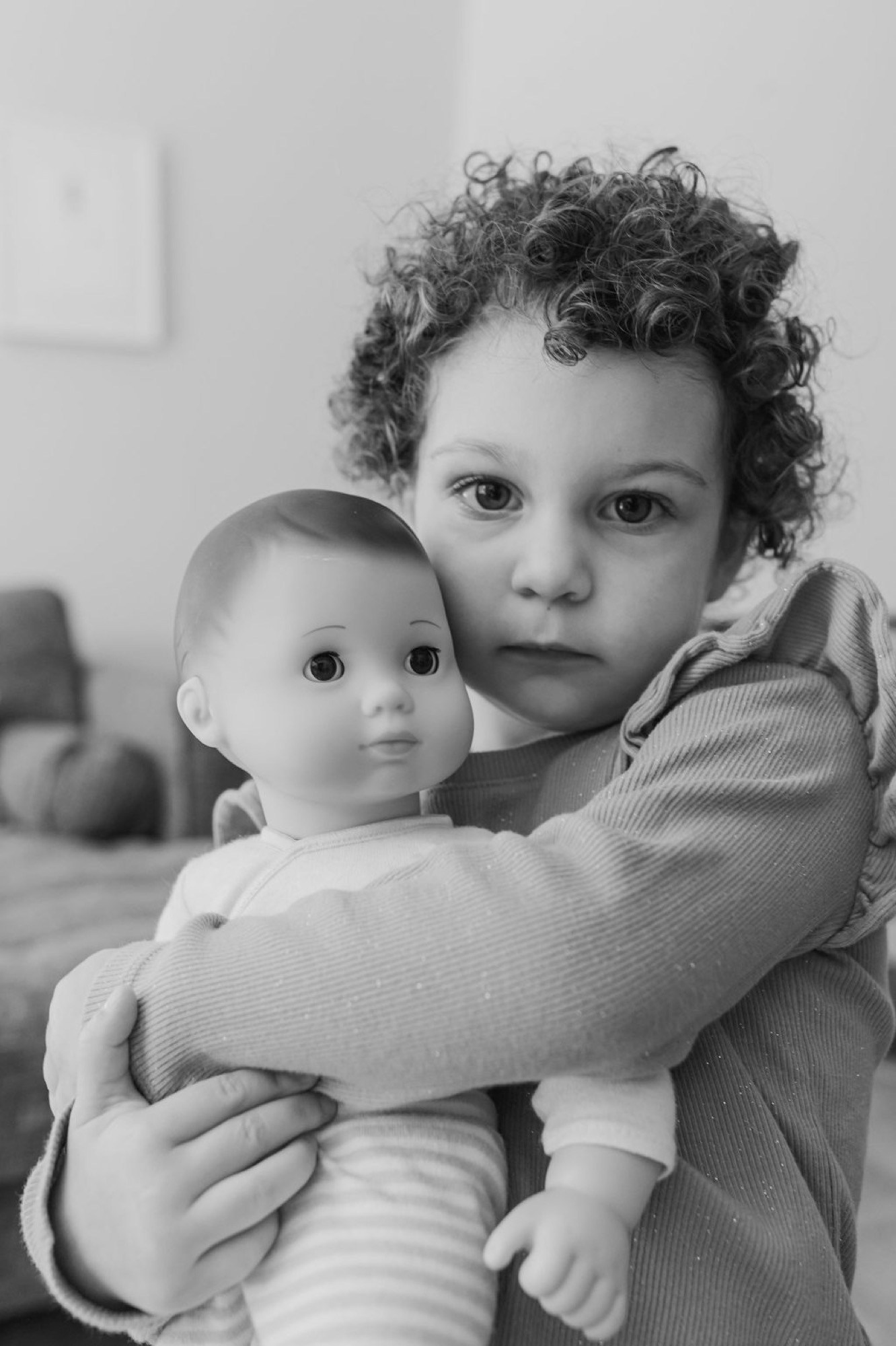 Child with curly dark hair holds a doll while gazing directly at camera with serious expression in soft, diffused indoor light
