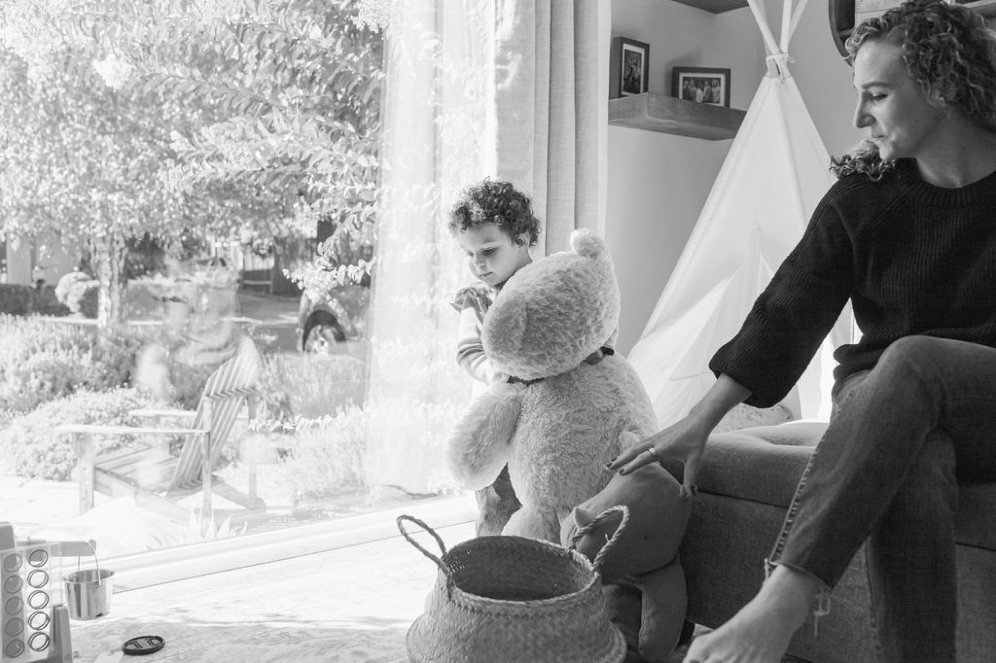 A mother guides her young child's hand toward a woven basket while a teddy bear sits between them near a sunlit window