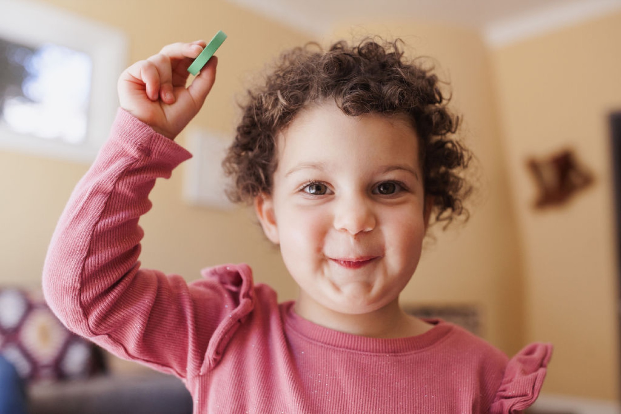 Curly-haired toddler in pink ribbed shirt raises arm triumphantly while holding a green crayon, beaming at camera indoors