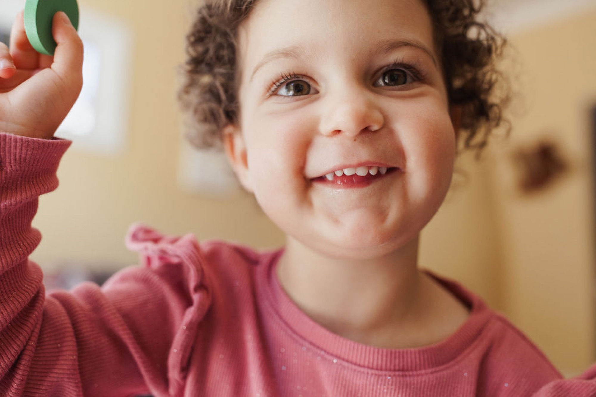 Toddler in burgundy shirt grins at camera with raised hand, curly hair catching warm indoor light in soft focus home setting