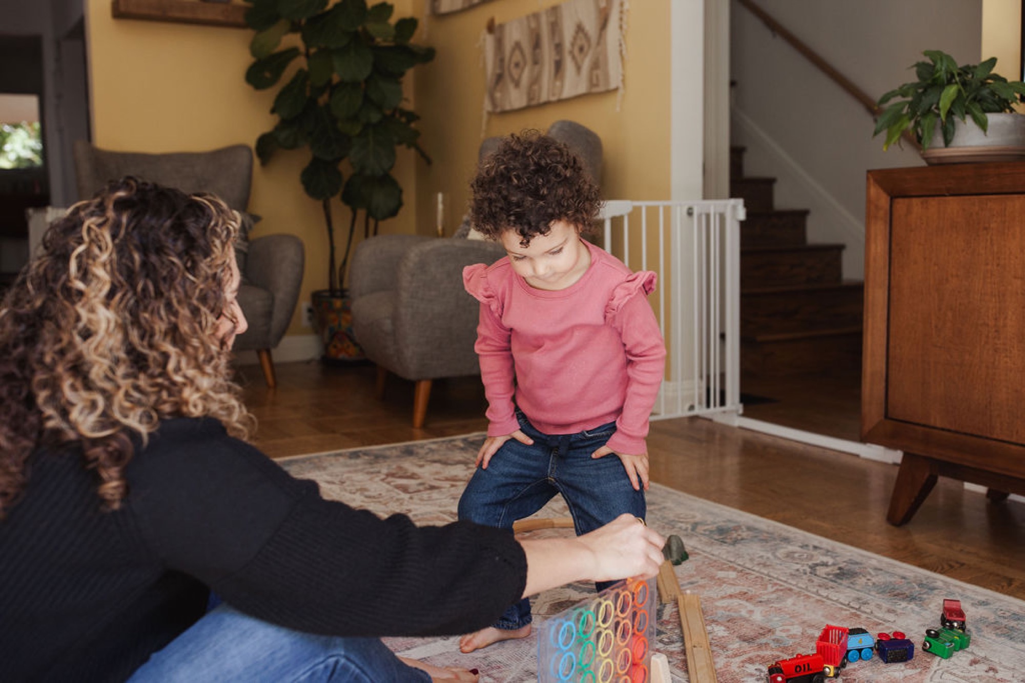 A toddler in a pink ruffled shirt gazes down at wooden toys while seated on a patterned rug, engaged with a patient caregiver in warm natural light