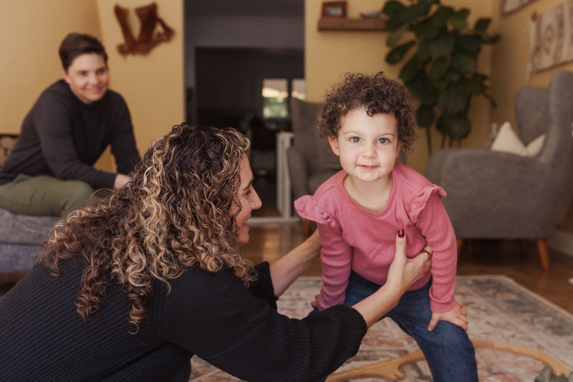 A woman with curly highlighted hair kneels to engage with a toddler in a pink striped shirt, while warm natural light fills the lived-in living room