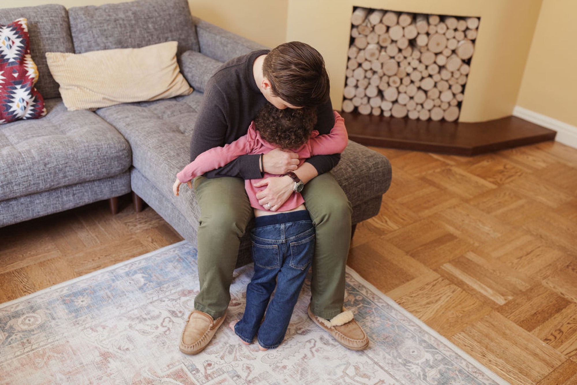 A parent holds a toddler close in a living room embrace, their foreheads nearly touching in a tender moment of connection