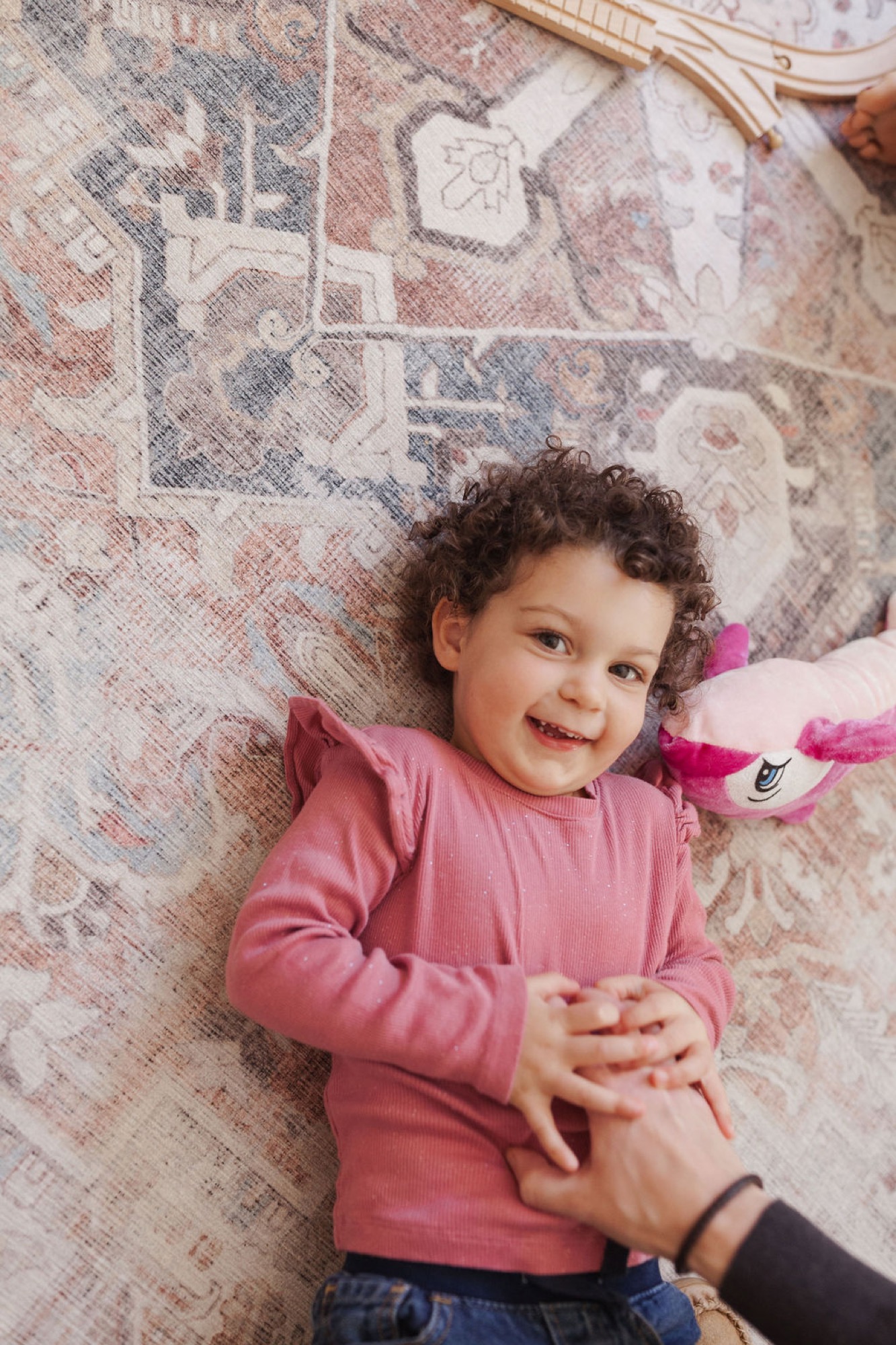 Curly-haired toddler in pink ribbed shirt laughs joyfully while lying on a patterned rug, holding an adult's hand