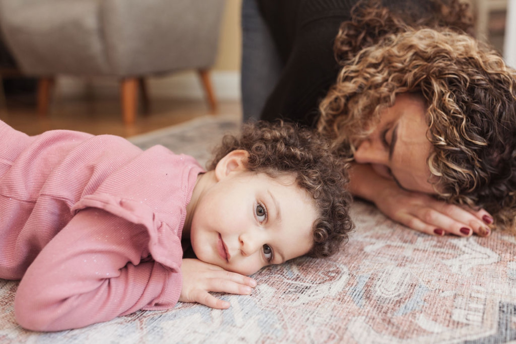 A mother and young child with curly brown hair lie on a textured rug, the child gazing at the camera while the mother tenderly kisses their head