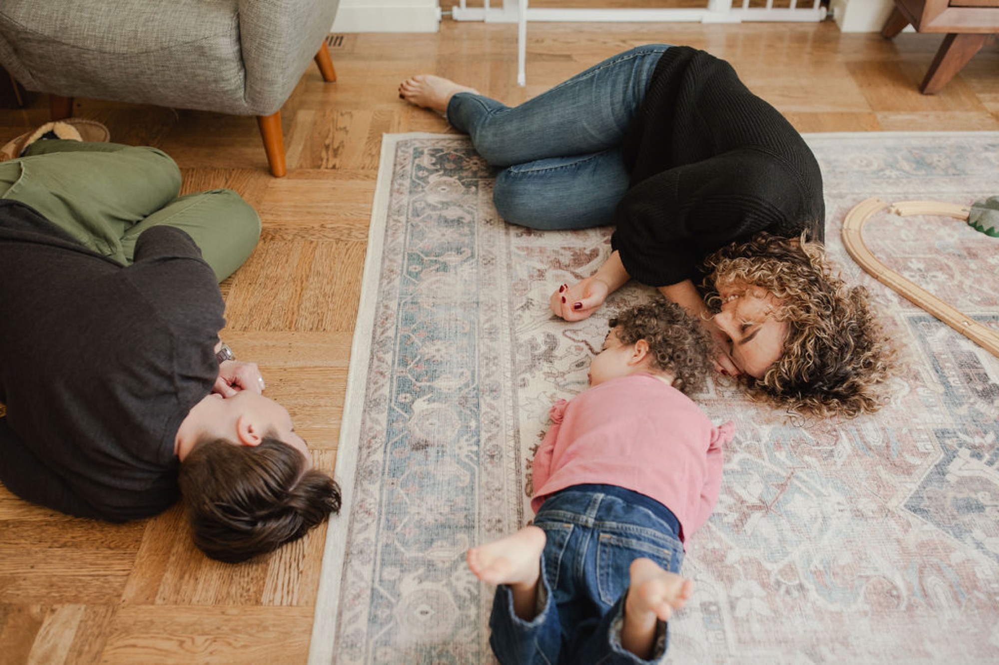 Three people lie sprawled on a patterned rug, heads tilted back in relaxed laughter, wooden floors and furniture visible around them