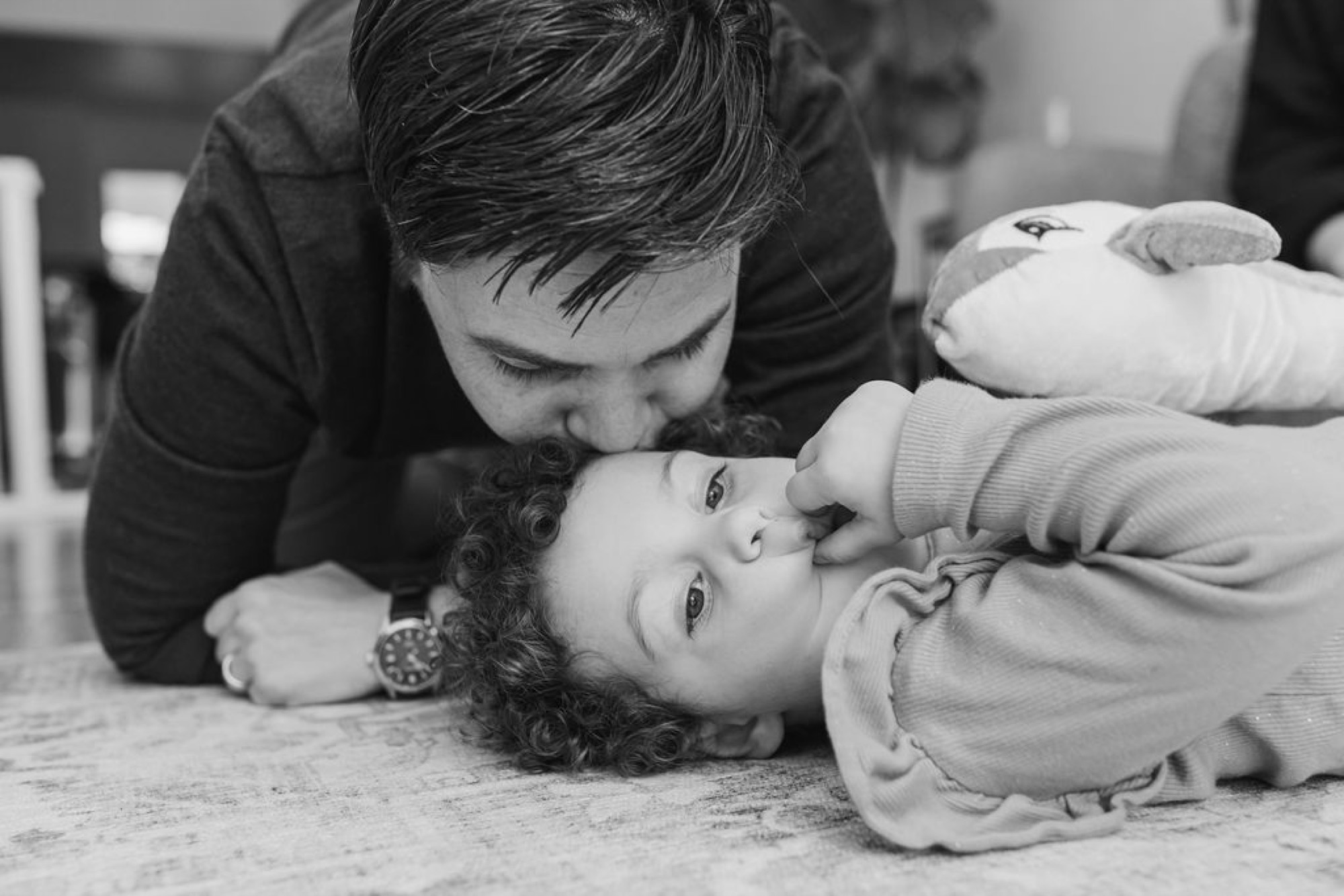 A parent and child lie face-to-face on a wooden floor, sharing an intimate moment of play and connection in soft natural light