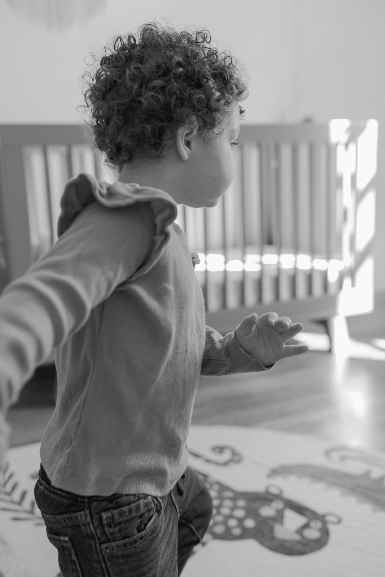 Toddler with curly hair stands barefoot in profile, arms outstretched in pure joy as bright window light streams across the nursery floor