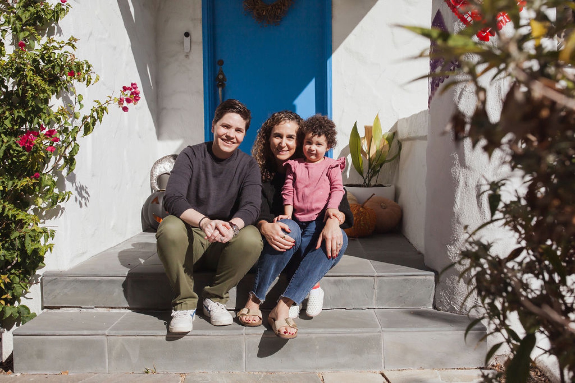 Three family members smile warmly on sun-drenched steps framed by flowering vines and white-washed walls with a bright blue door