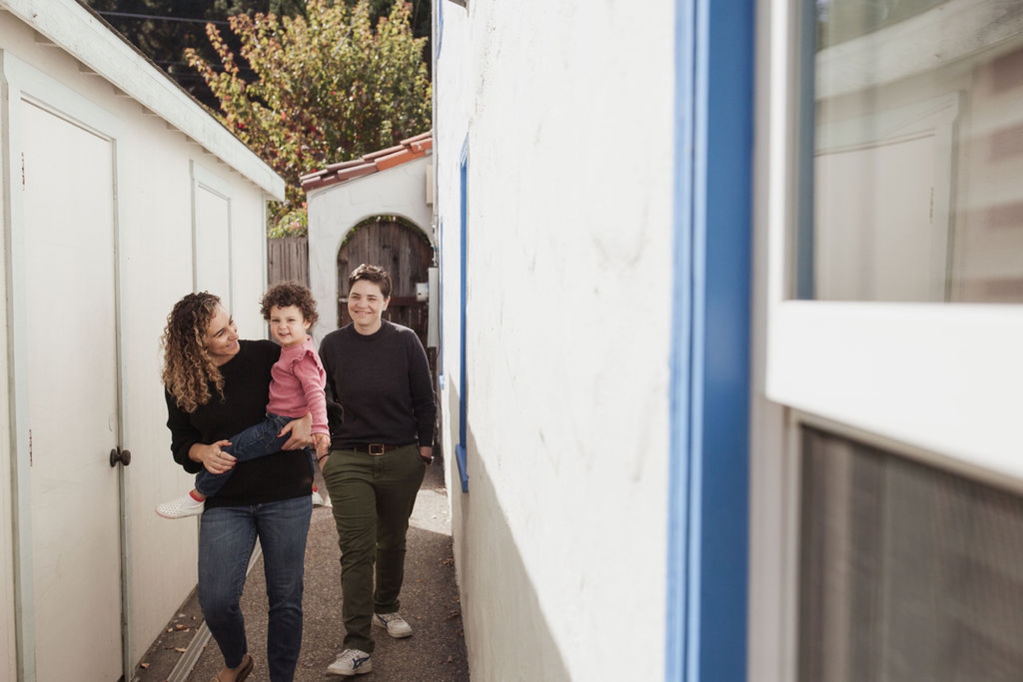 A mother holds her toddler while walking alongside an older child through a narrow white-walled passageway, all three smiling naturally toward the camera