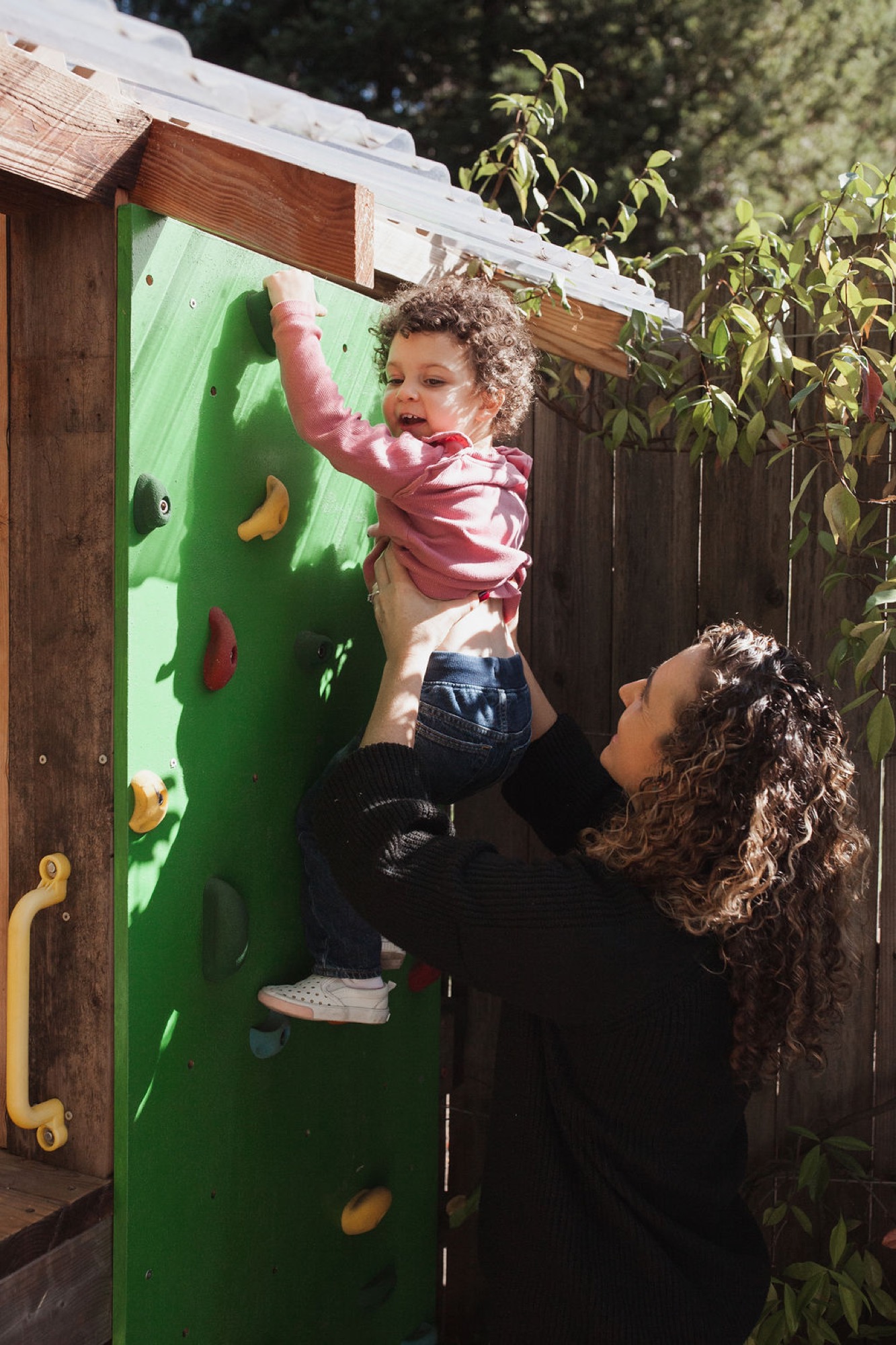 A caregiver lifts a toddler toward colorful climbing holds on a bright green play wall, capturing genuine joy and physical engagement in dappled sunlight