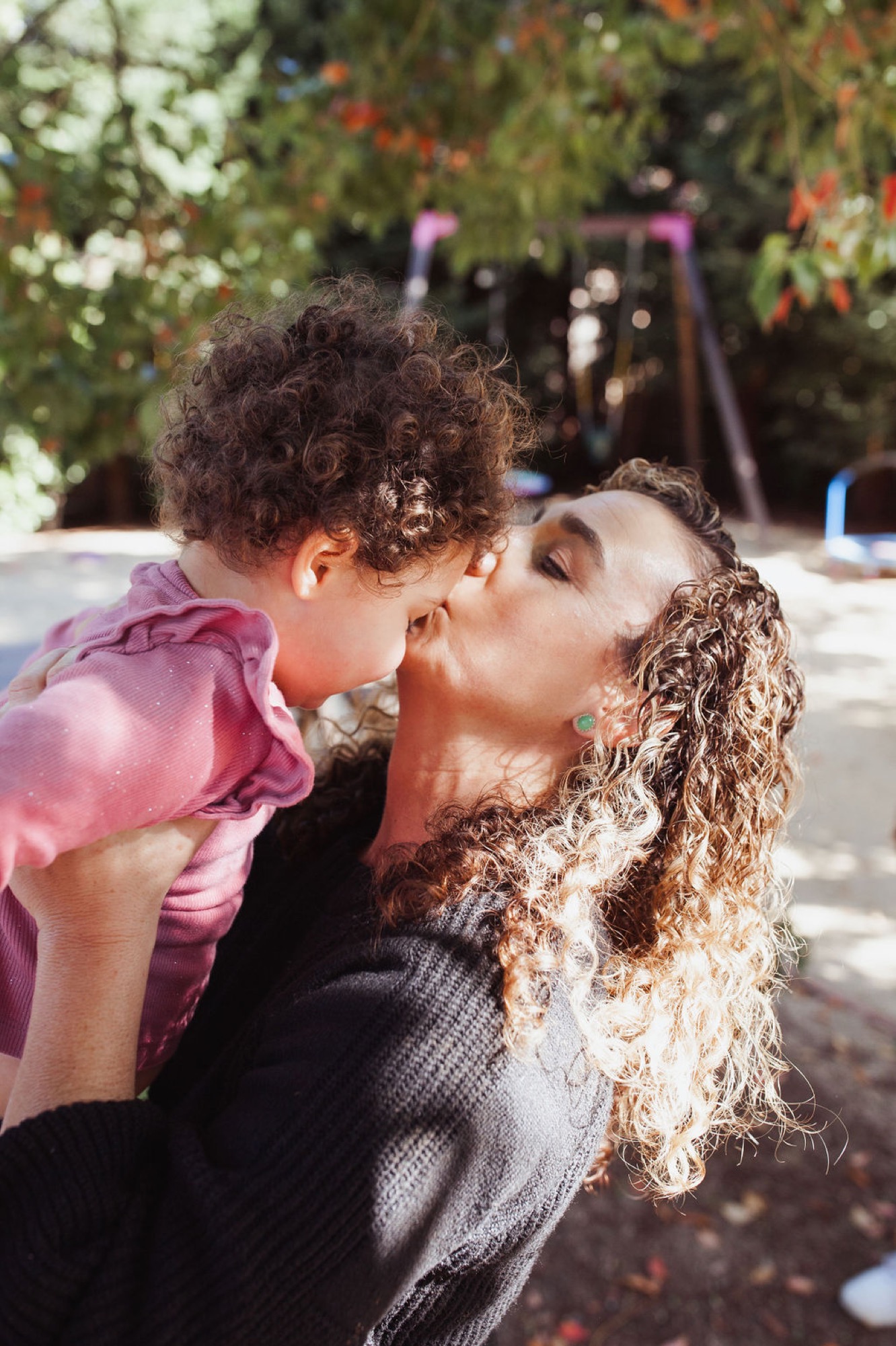 A mother kisses her toddler's cheek in bright sunlight while holding him close, with lush green foliage and garden structures softly blurred behind them