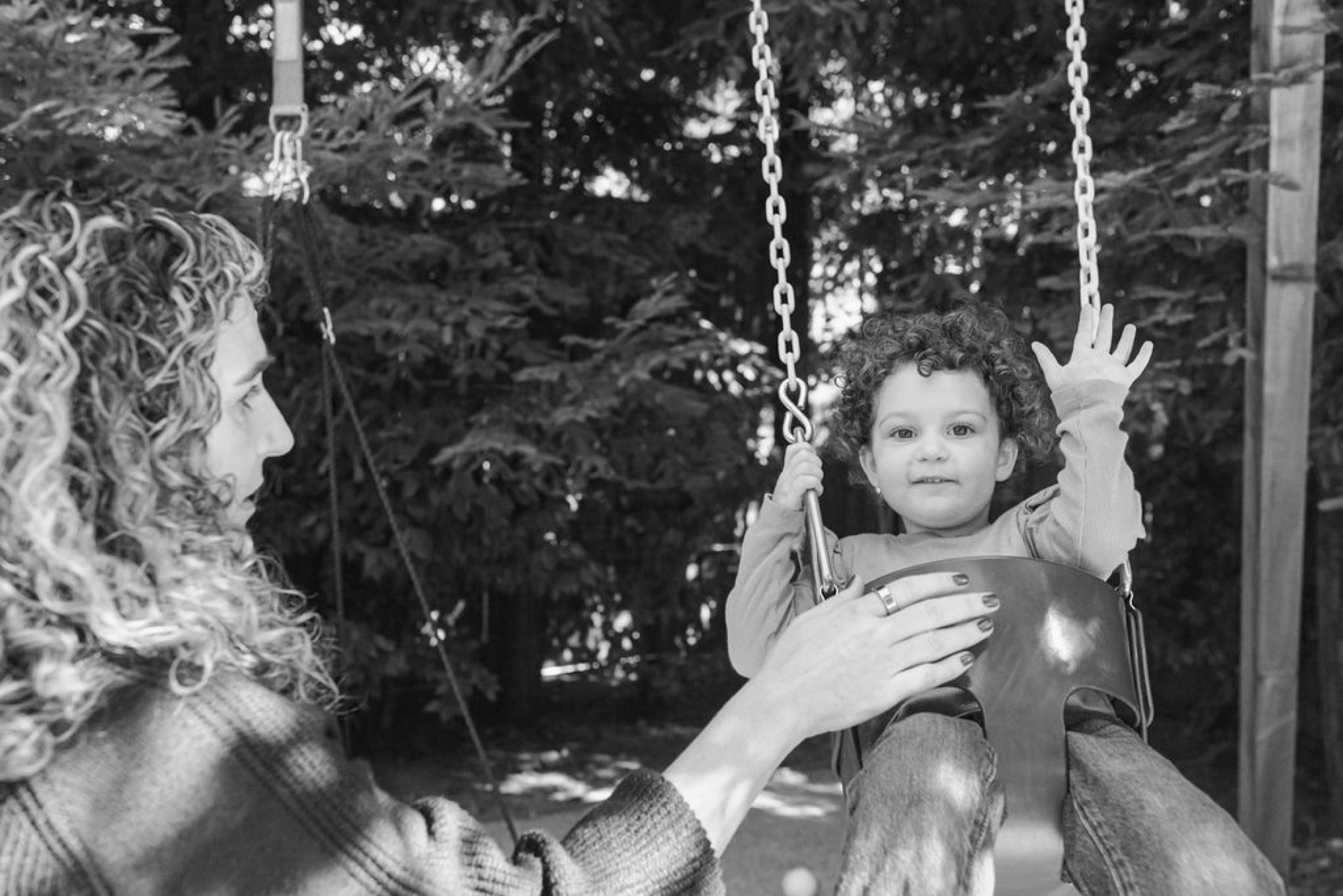 A woman pushes her curly-haired toddler on a swing, his hand raised with joy as dappled sunlight filters through trees behind them