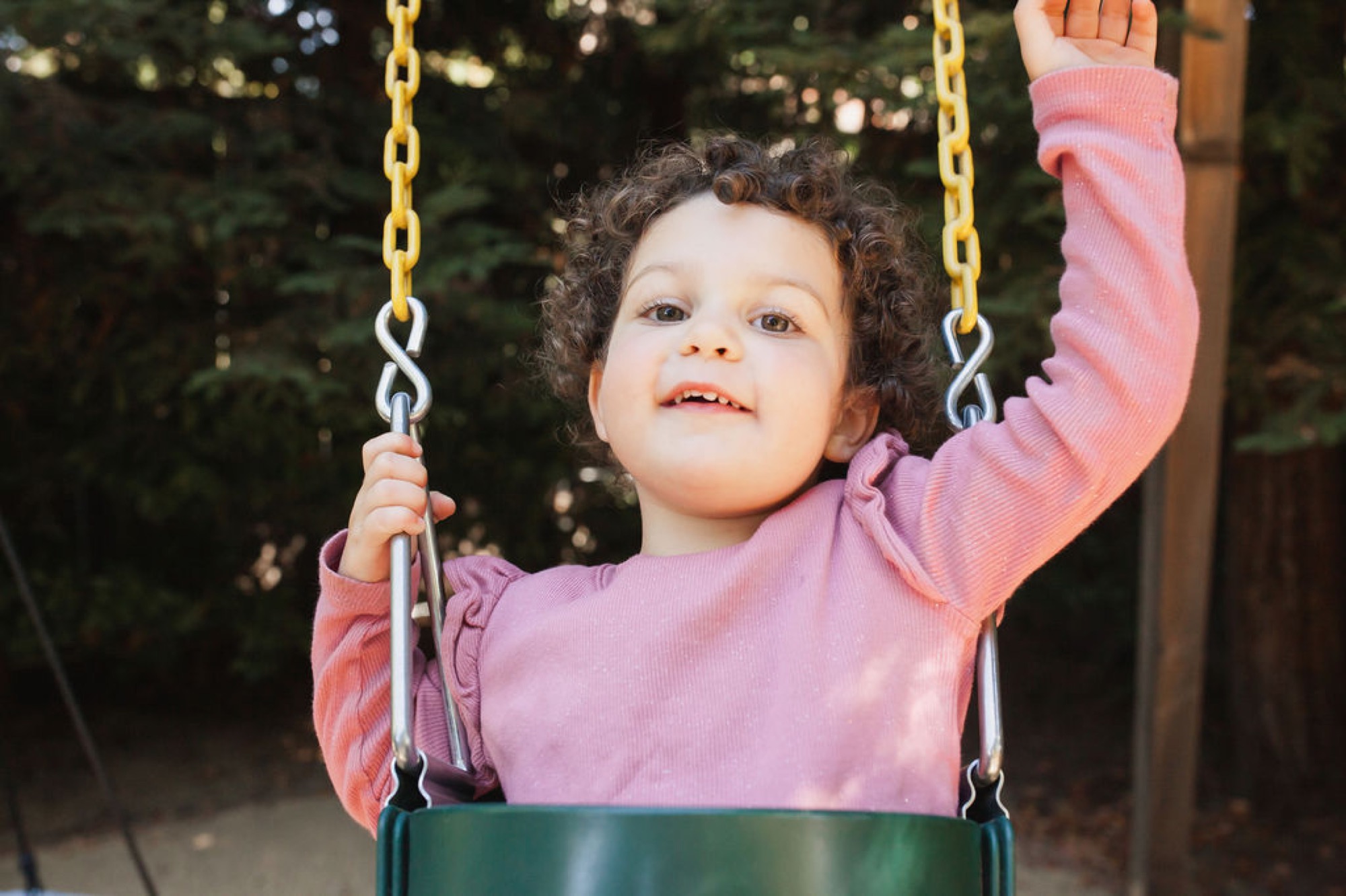 A toddler with curly dark hair grins widely while swinging, one arm raised joyfully in a pink ribbed shirt against blurred green foliage