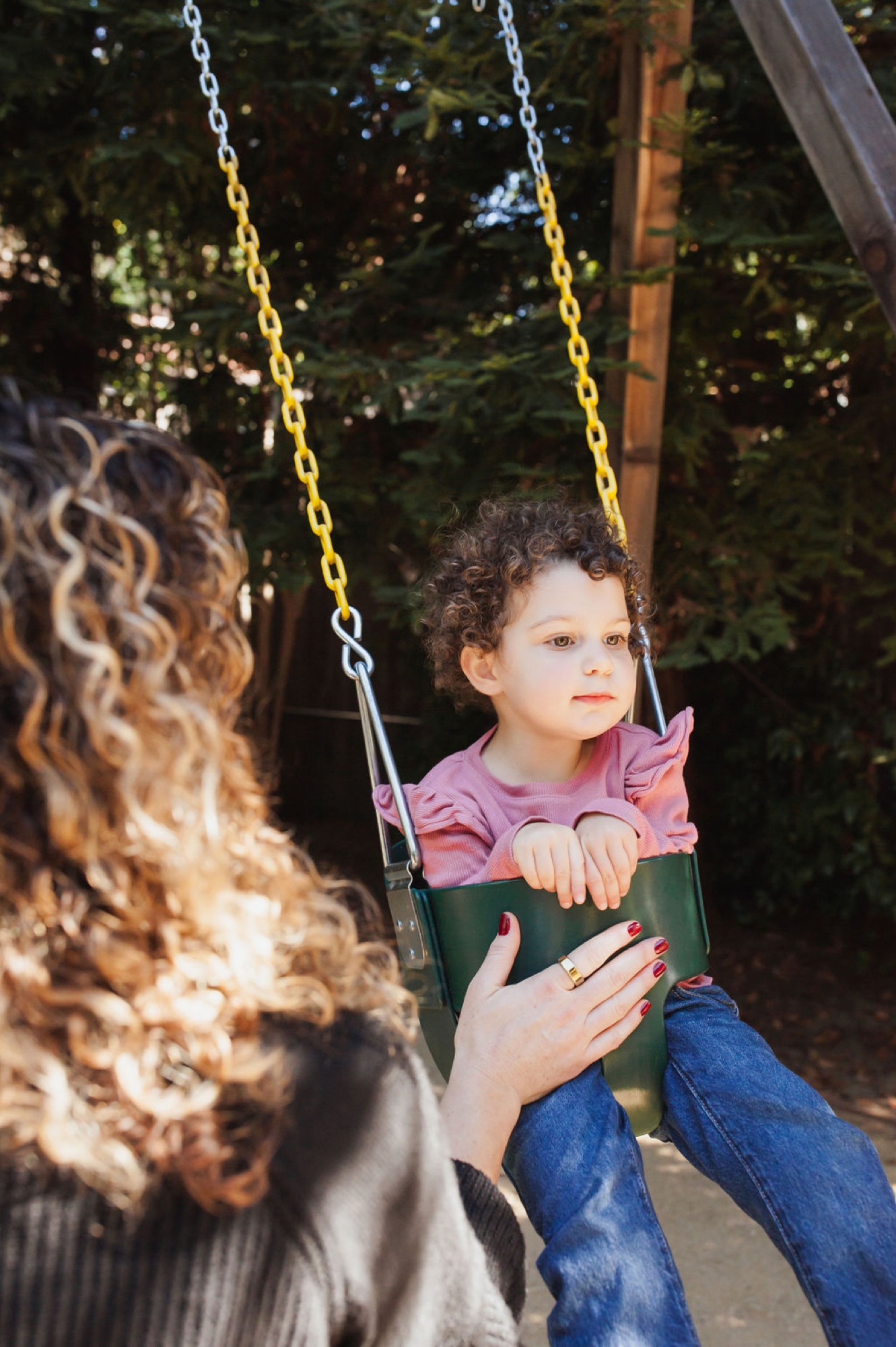 A caregiver gently pushes a curly-haired toddler on a green swing, dappled sunlight filtering through lush trees behind them