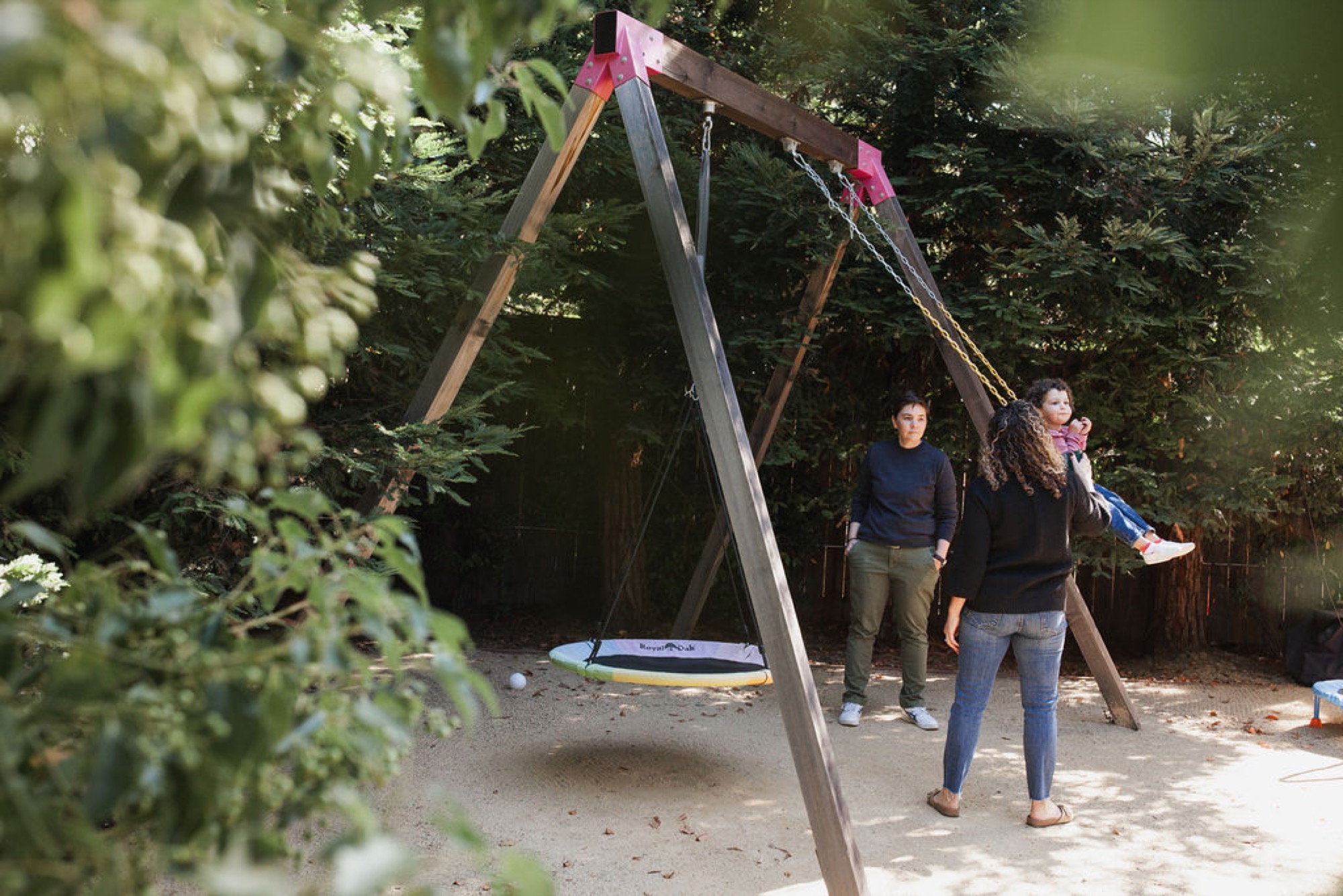 Three children stand beneath a wooden swing set nestled in dappled forest light, with one girl mid-stride and natural greenery framing the scene