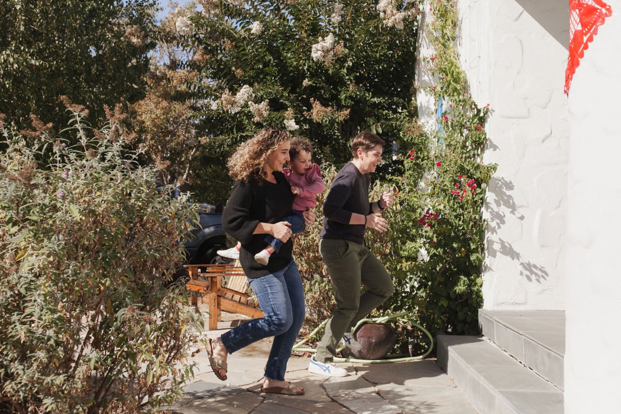 A woman carrying a toddler jogs alongside a boy through a narrow vine-covered garden pathway, dappled sunlight creating dynamic shadows on the white wall