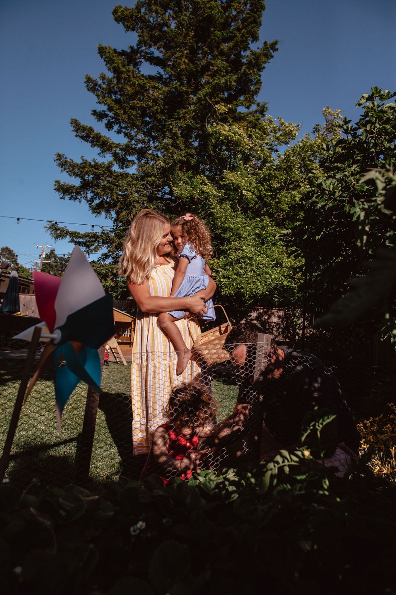 A blonde woman in a striped dress holds her curly-haired daughter, both smiling gently in dappled sunlight beneath towering oak trees