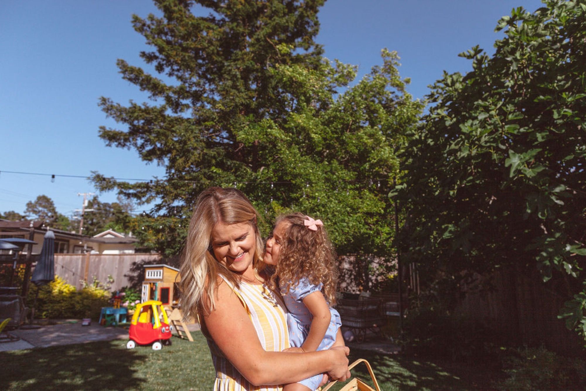 A mother holds her young daughter close in a sunlit backyard, both smiling warmly as the girl whispers in her ear