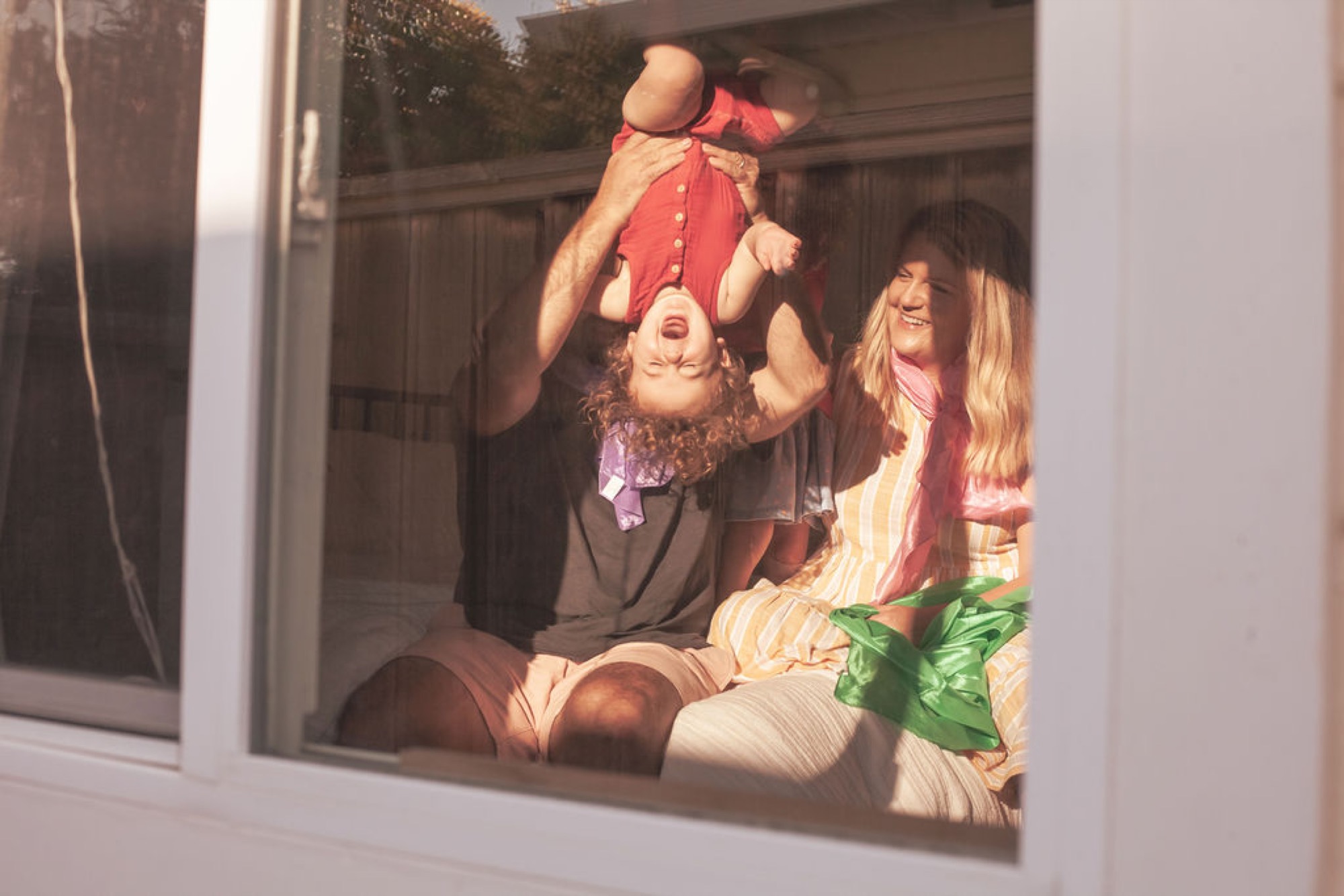 Father lifts laughing infant overhead while mother sits beside them, golden afternoon light streaming through the window onto their joyful faces