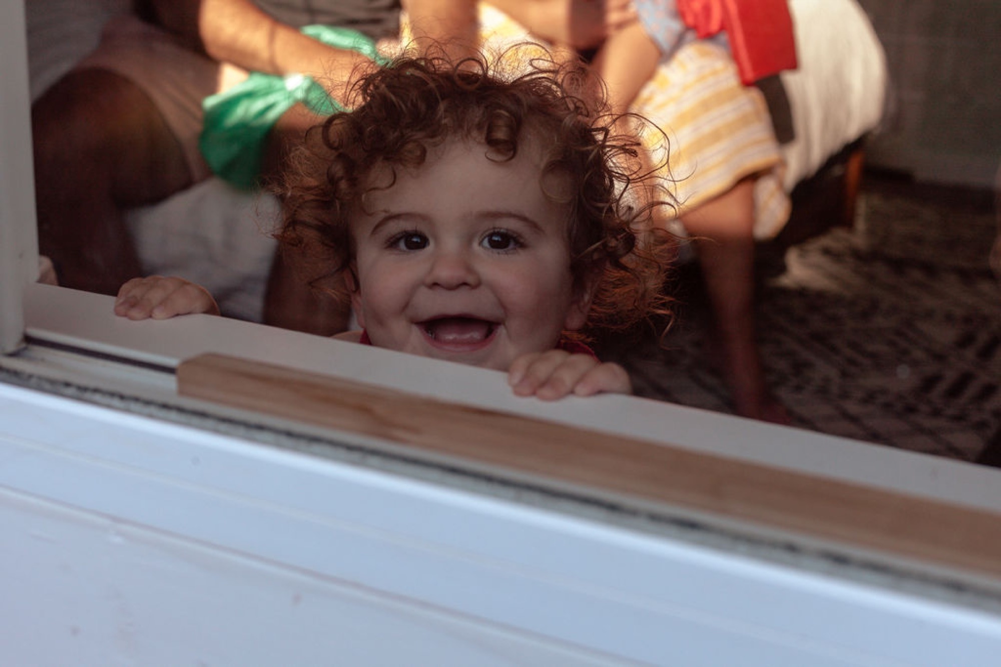 A curly-haired toddler grins widely at the window, bathed in warm afternoon sunlight