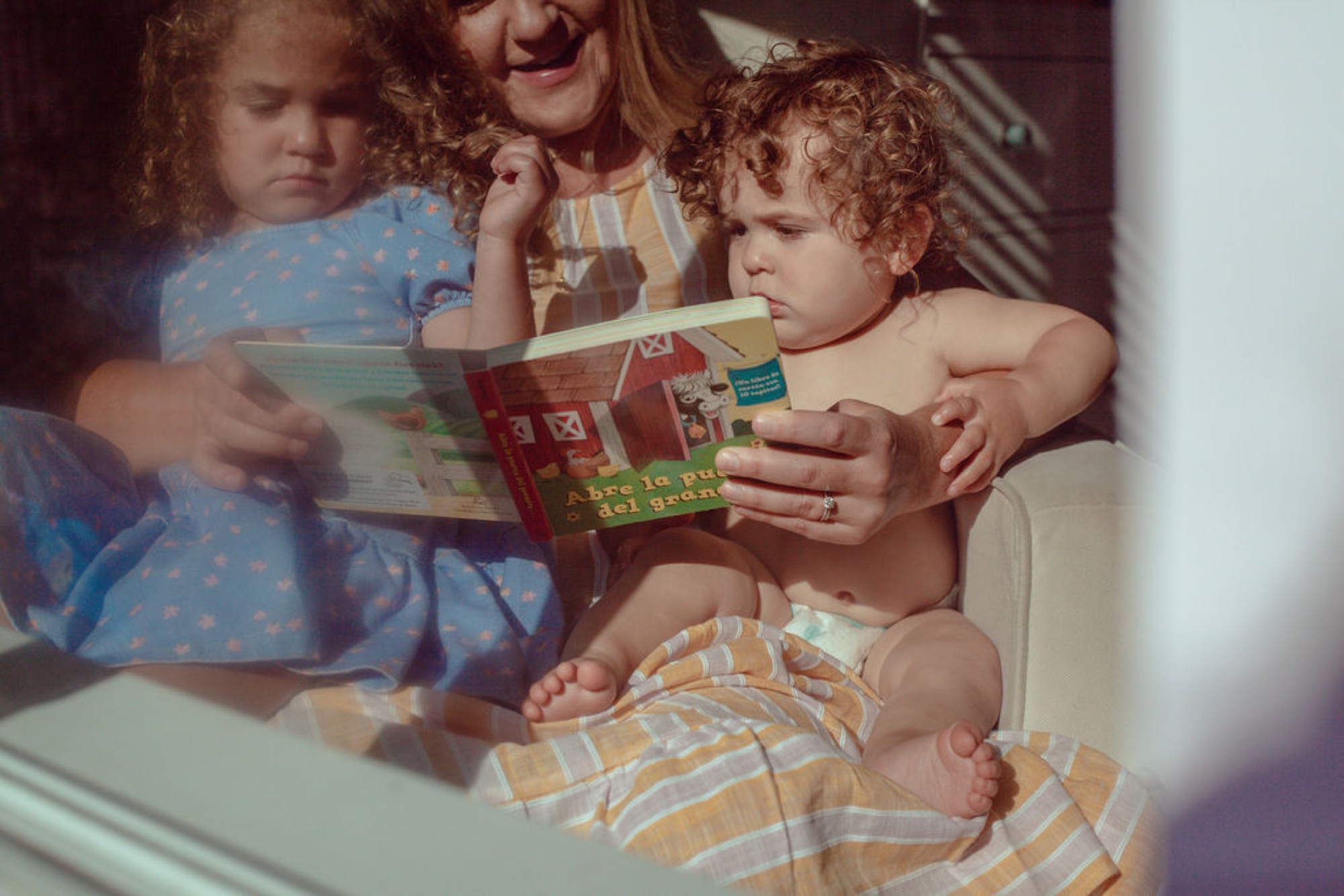 A woman reads a Spanish-language picture book to two curly-haired toddlers in warm afternoon sunlight streaming across their laps