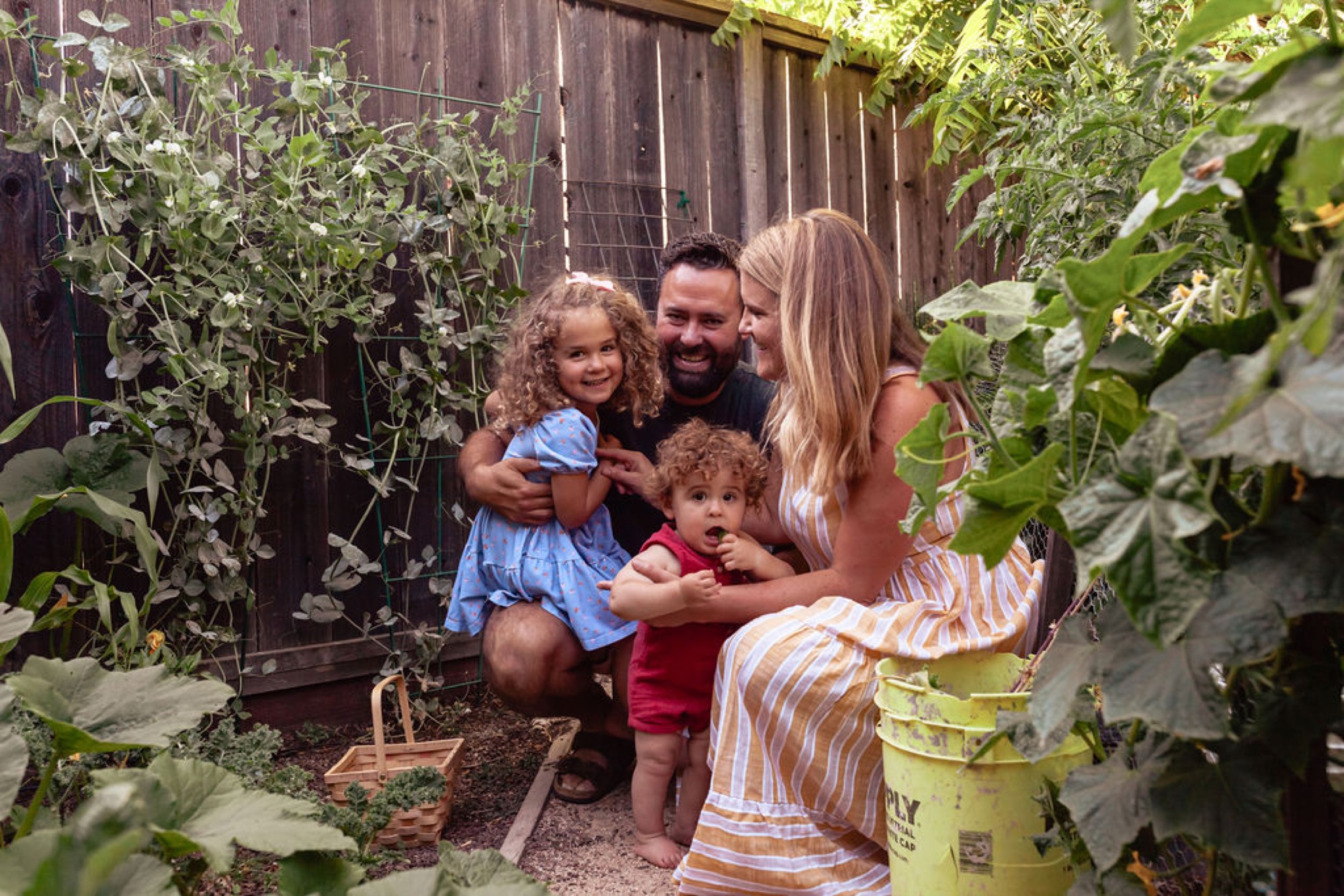 Family of four smiles among lush garden plants, surrounded by climbing vines and flowering tomato plants in afternoon light