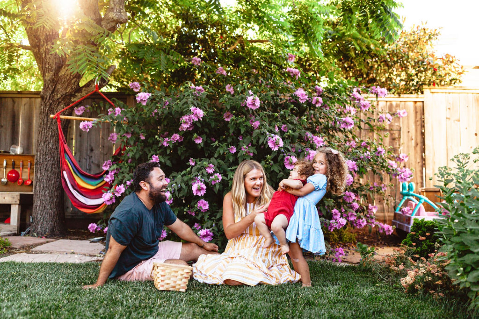 A family of four sits on grass beneath blooming pink flowers, the father and mother smiling while holding their two young children in warm afternoon sunlight