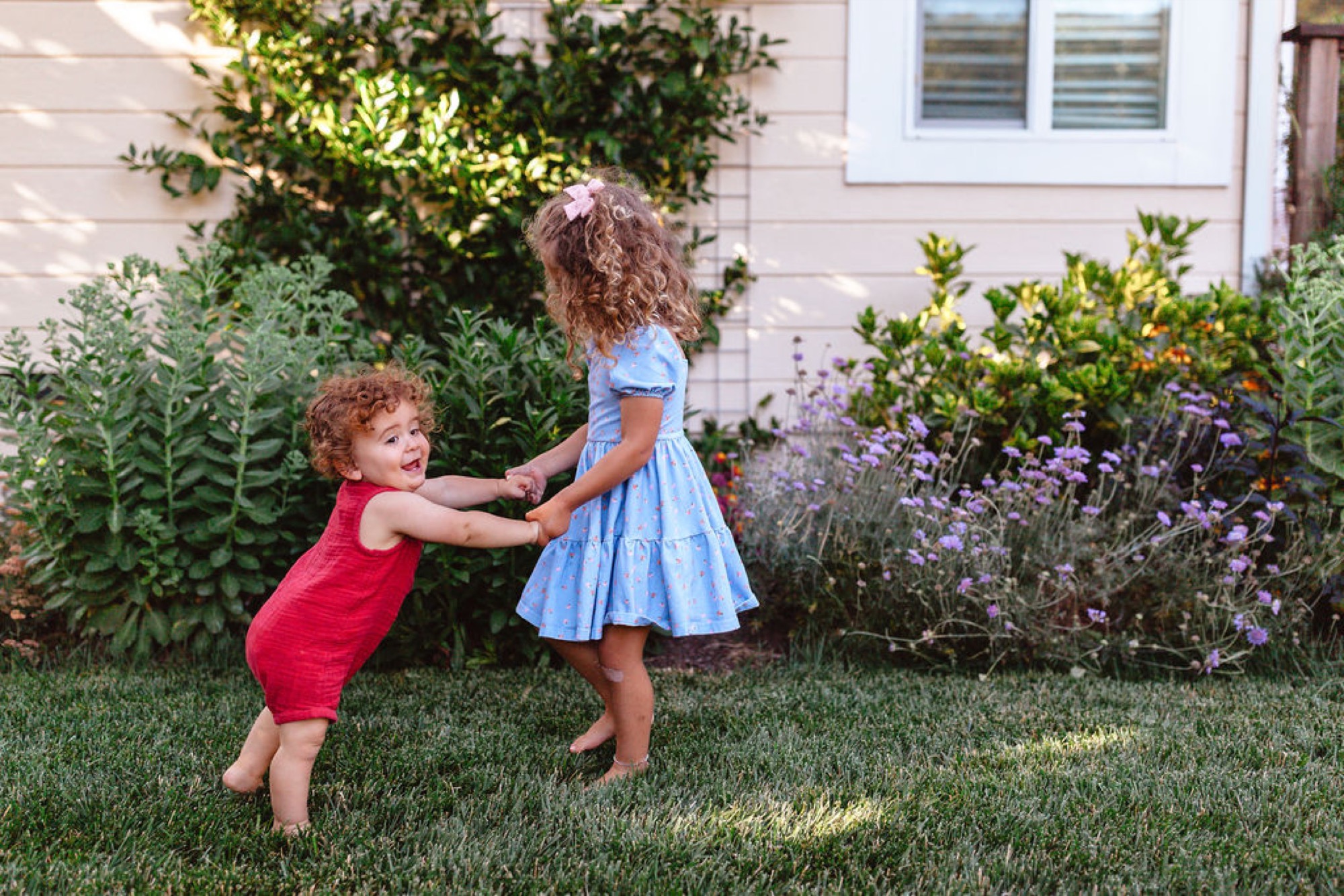 Two toddlers with curly hair hold hands while playing in a sunlit backyard garden