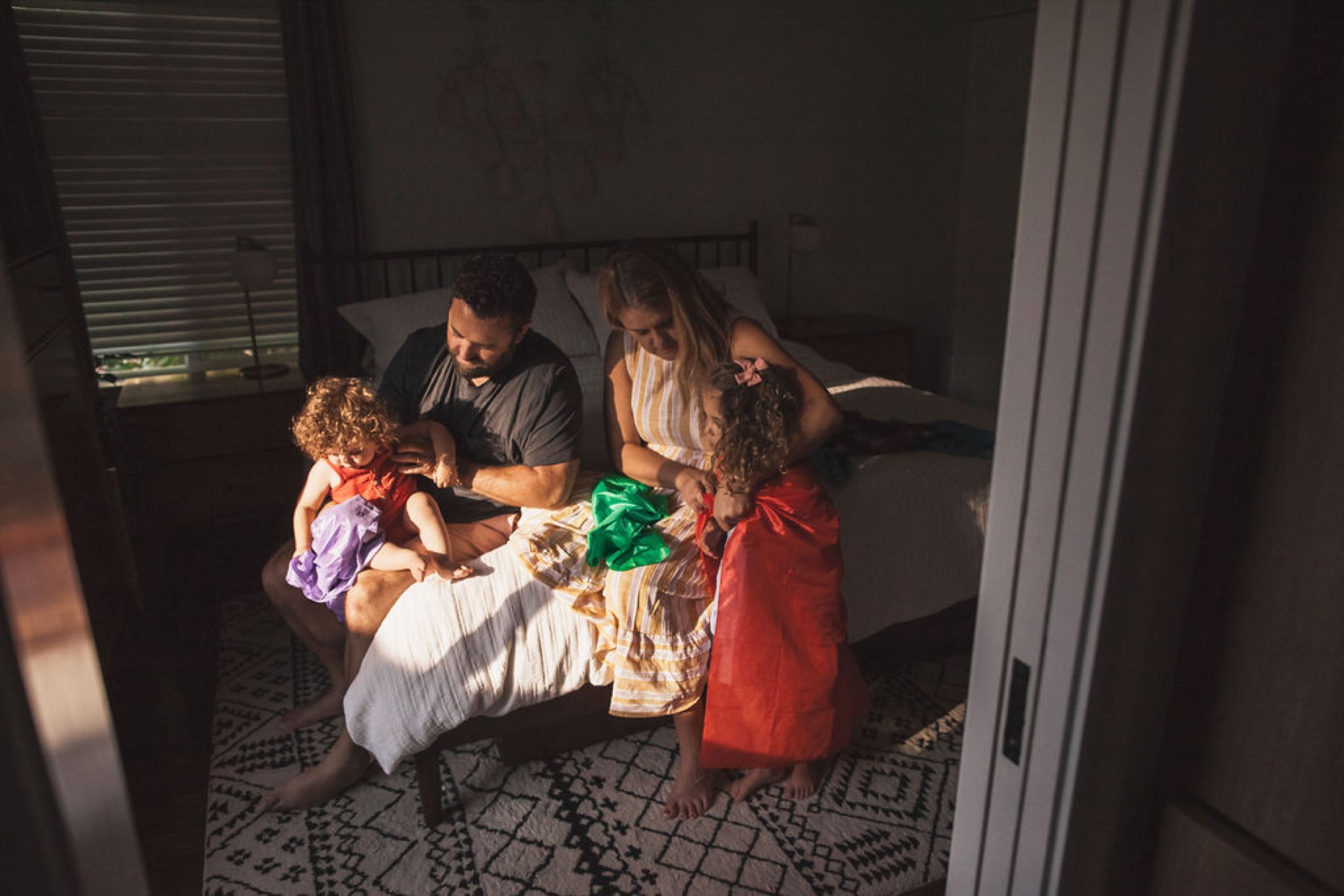 Parents and two young children sit on a bed in golden afternoon light, opening wrapped gifts together with genuine joy and engagement