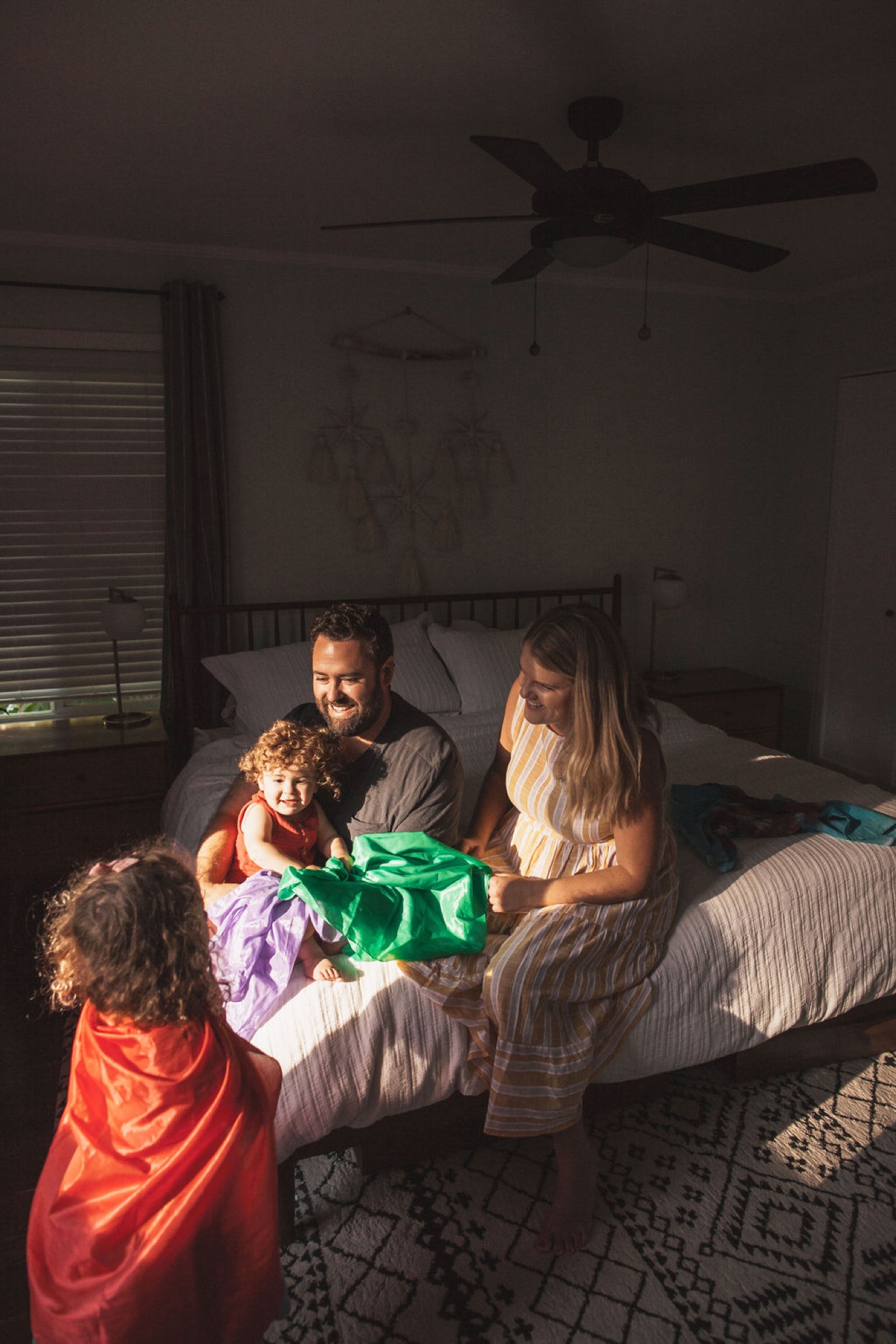 A father and mother sit on their bed with two young children wearing colorful capes, bathed in warm natural sunlight streaming through the window