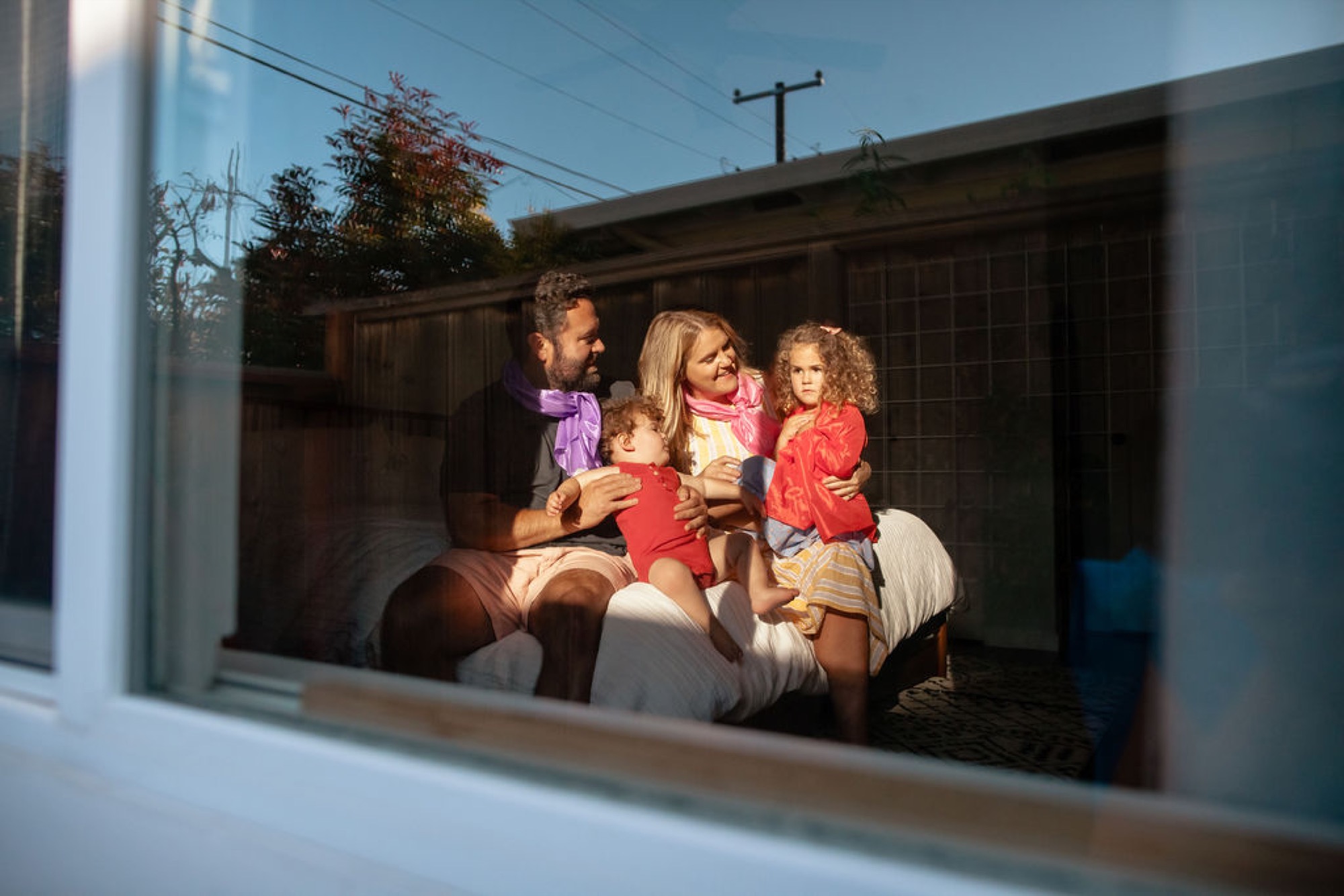 A family of four sits together on a white bench in their sunny backyard, children in bright red