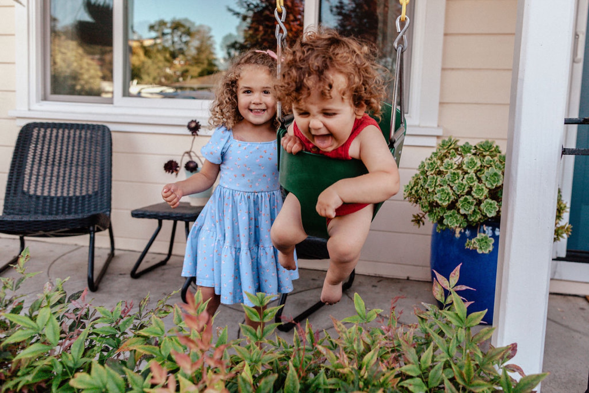 Two curly-haired toddlers laugh joyfully on a porch swing, with one in a blue dress and one in red, bathed in warm afternoon sunlight
