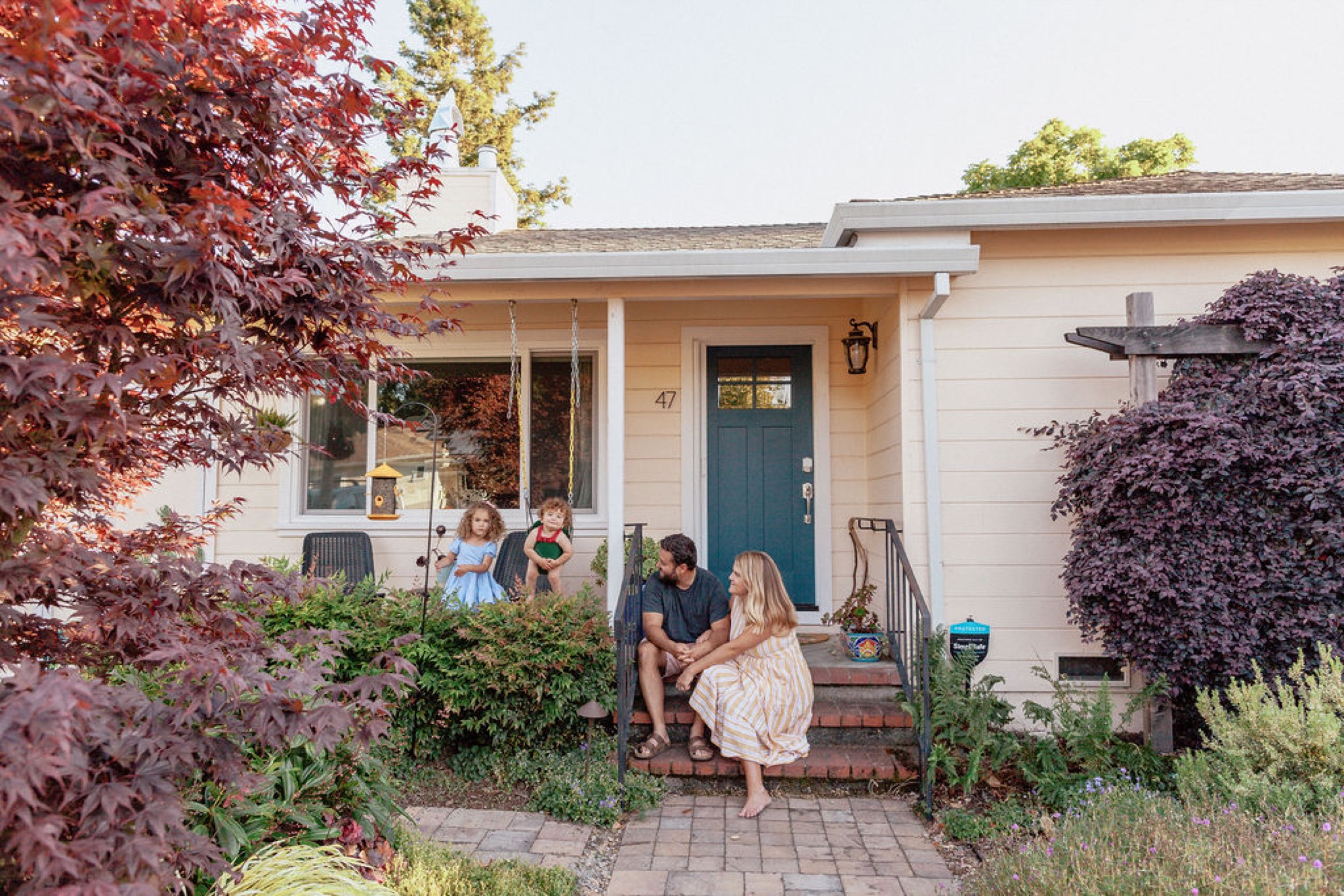 A family sits together on brick front steps, framed by a deep red Japanese maple and purple-leafed vines