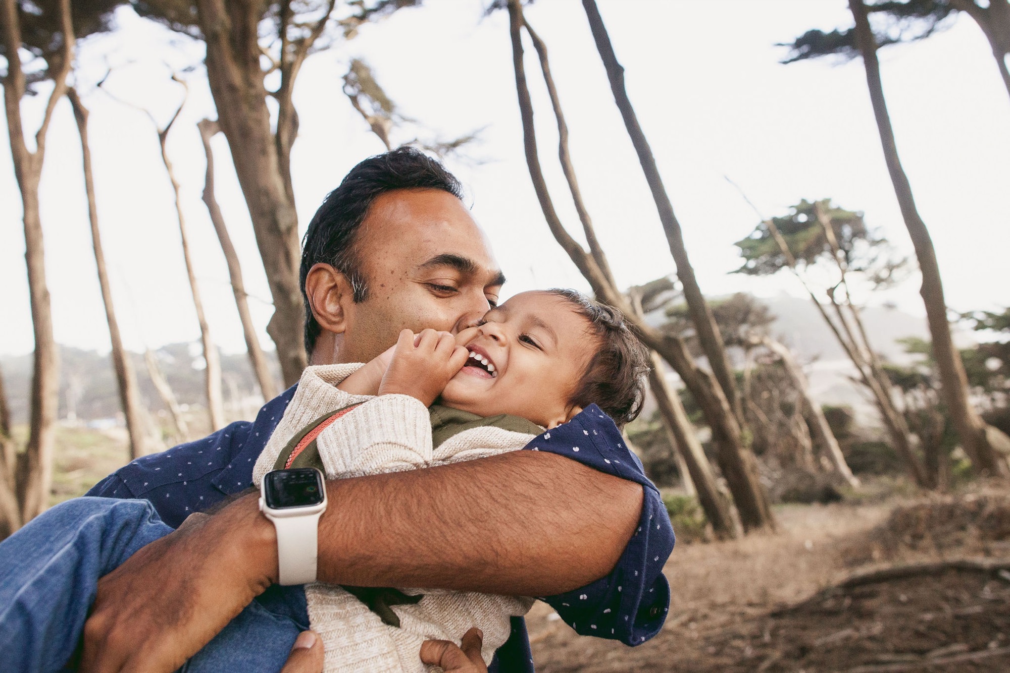 A father holds his laughing toddler among towering Monterey cypress trees, kissing her cheek as golden afternoon light filters through the canopy