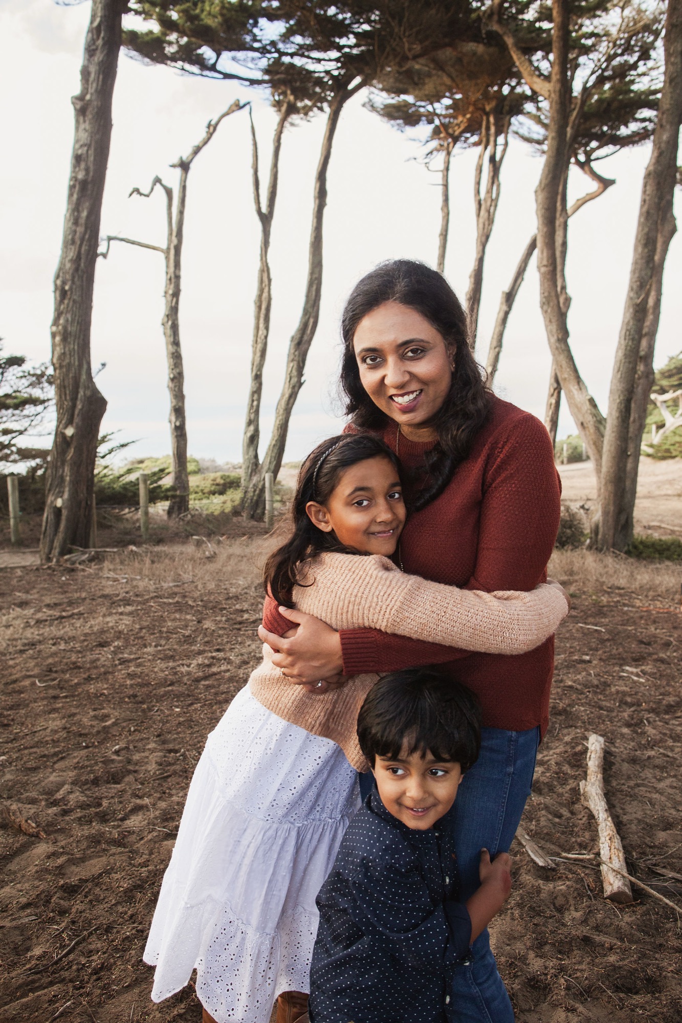 A mother embraces her two smiling children among tall, wind-sculpted cypress trees with soft coastal light filtering through the canopy