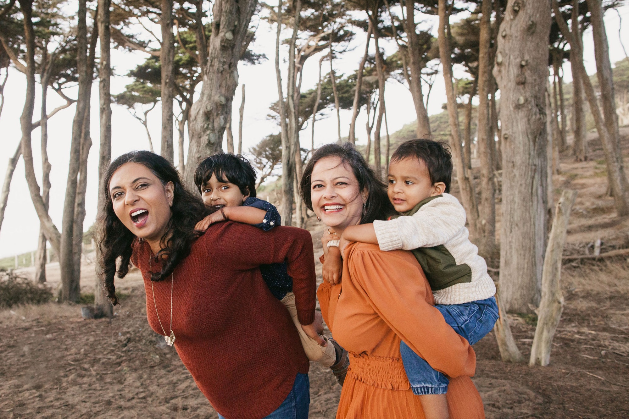 Two women carry laughing children piggyback through a towering cypress grove, their joyful expressions and warm clothing suggesting a breezy coastal afternoon